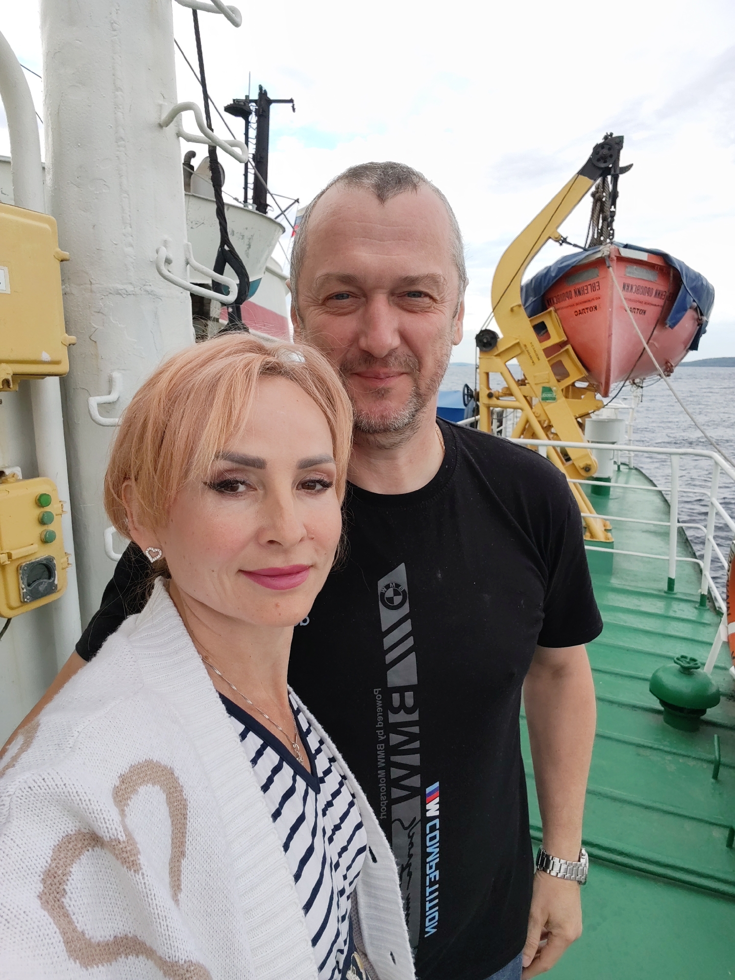 A medium shot shows a man and a woman standing on the deck of a boat. The woman is on the left, facing the camera with her head tilted slightly to the right. She has short, light blonde hair and is wearing a white cardigan with a heart pattern over a striped shirt. She has on heart-shaped earrings and a necklace. The man is on the right, also facing the camera. He has short, light brown hair and stubble, and is wearing a black t-shirt with a graphic design. Both individuals appear to be smiling. Behind them, the boat's railing and deck are visible, along with a yellow control panel on the left and a life raft suspended from a crane on the right. The water is visible in the background, with a cloudy sky above. The location is identified as the city of Shal'skii, Russia.