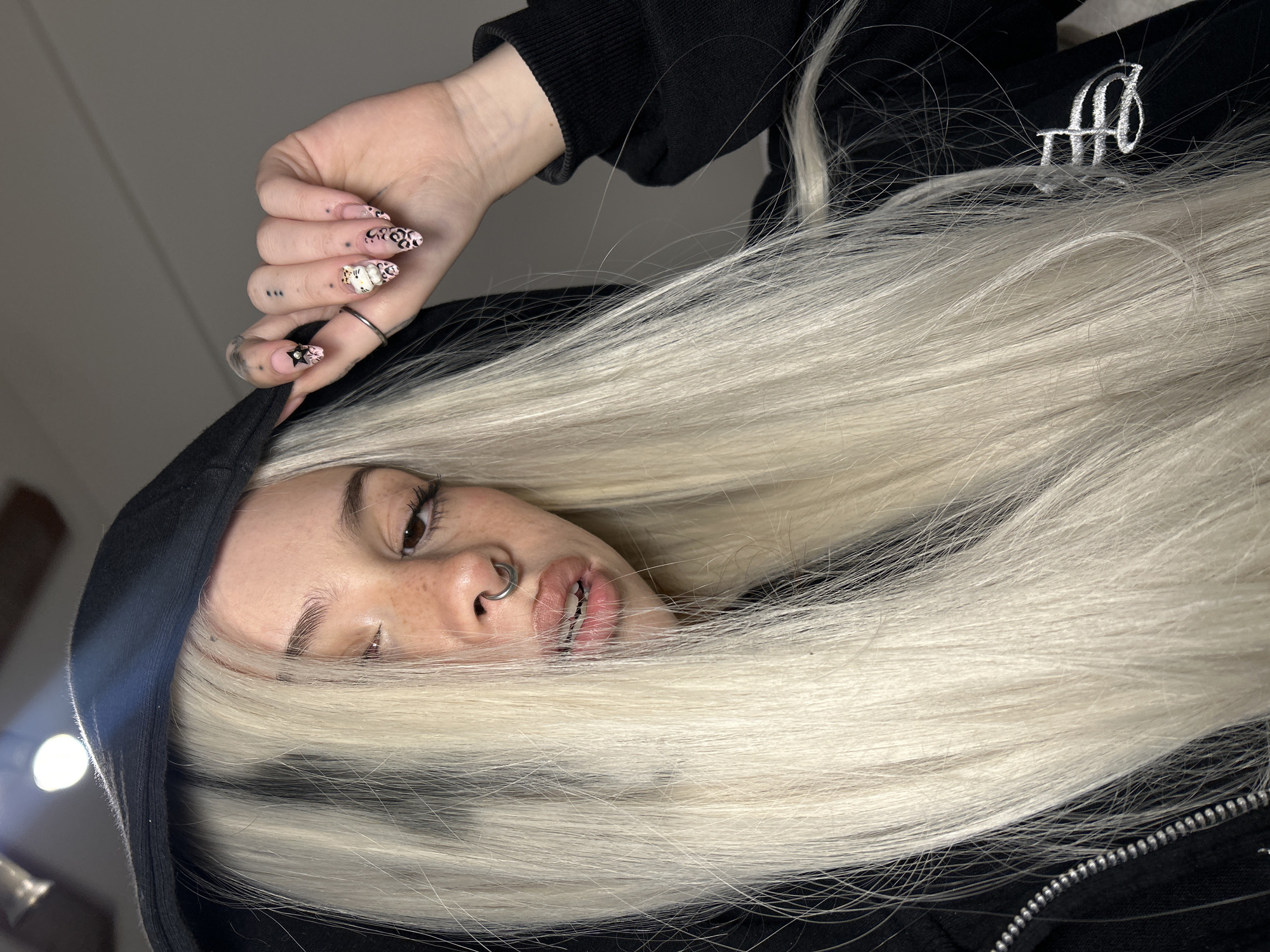 The image captures a close-up, overhead perspective of a person with distinct features, primarily their face and hand, framed by a voluminous cascade of platinum blonde hair. The individual is wearing a black hooded sweatshirt.
The person's face is positioned towards the bottom of the frame, featuring light skin, visible freckles on the nose and cheek area, and dark brown eyes that reflect a bright artificial light source, possibly a ring light. Their eyebrows are dark and shaped, and eyelashes appear enhanced. A prominent silver septum piercing is visible. The lips are slightly parted, revealing metal dental braces on the lower teeth.
A left hand is raised into the frame, with fingers slightly curled. The hand has several small, dark dot tattoos on the back and fingers, including a distinctive pattern of three dots. The nails are long and almond-shaped, adorned with elaborate designs: pink leopard print, a white character resembling Hello Kitty with a pink bow, and a black star accompanied by pink floral elements. A simple silver ring is worn on the ring finger.
The long, straight, bright blonde hair spreads widely, occupying a significant portion of the image and covering much of the background. The person is dressed in a black hoodie, part of which features a white, stylized, calligraphic logo that appears to be two intertwined letters, possibly "AH" or "HH," embroidered on the fabric. A silver zipper is also visible on the hoodie.
The setting appears to be indoors, indicated by the artificial lighting. There are no specific visual cues within the image to directly confirm the location as Buenos Aires, Argentina. The overall activity is a posed photograph, with the subject looking towards the camera from an unconventional angle, contributing to a distinctive and stylized aesthetic.