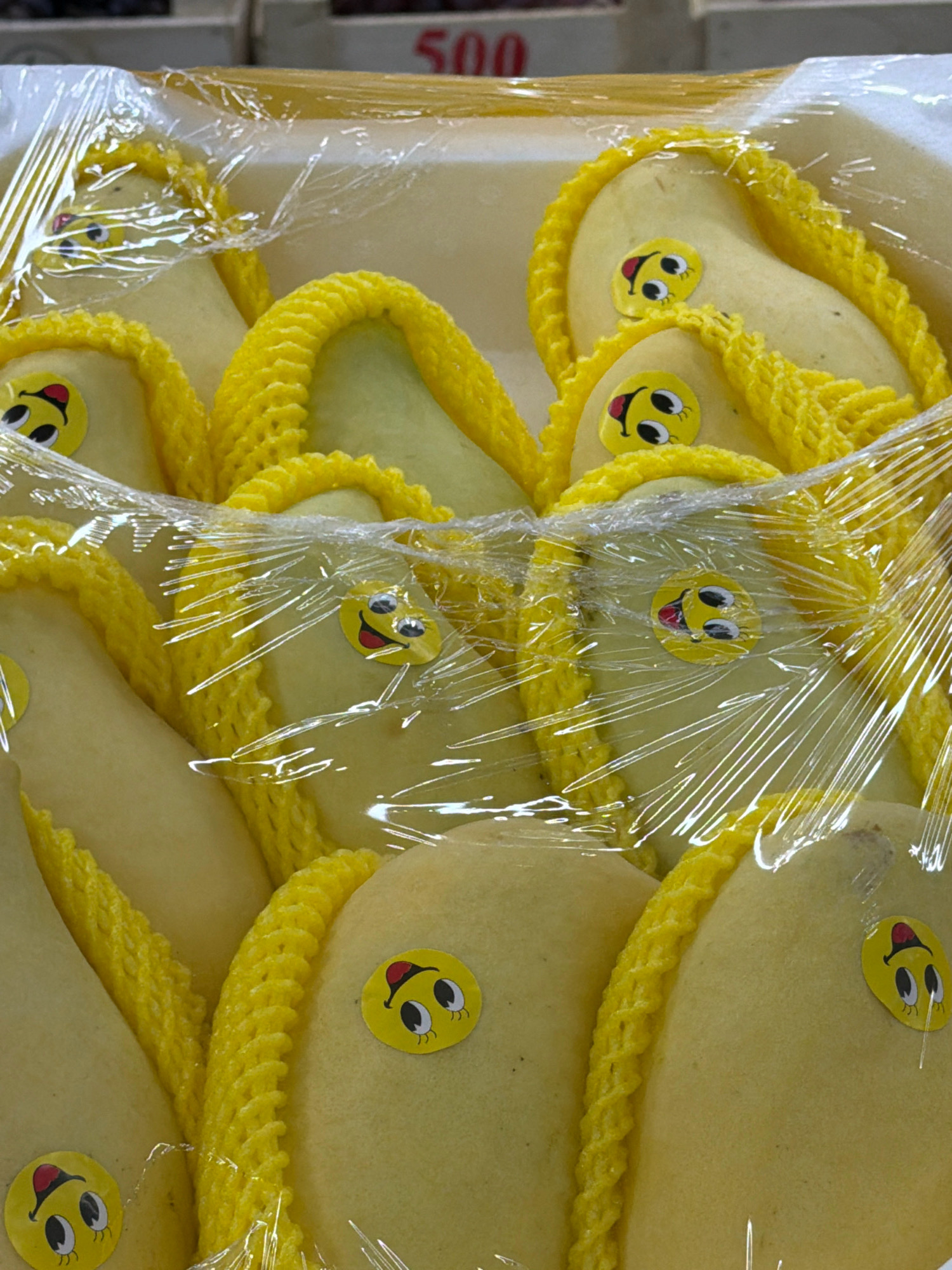 The image shows several yellow fruits, likely pears, individually wrapped in clear plastic and cushioned by yellow foam netting. Many of the fruits have circular stickers with cartoon faces depicting surprised expressions with wide eyes and red mouths. The fruits are arranged in a container, with a price tag reading "500" visible in the background. The scene appears to be a retail display in a market or grocery store. The location is not specified in the image or accompanying text.