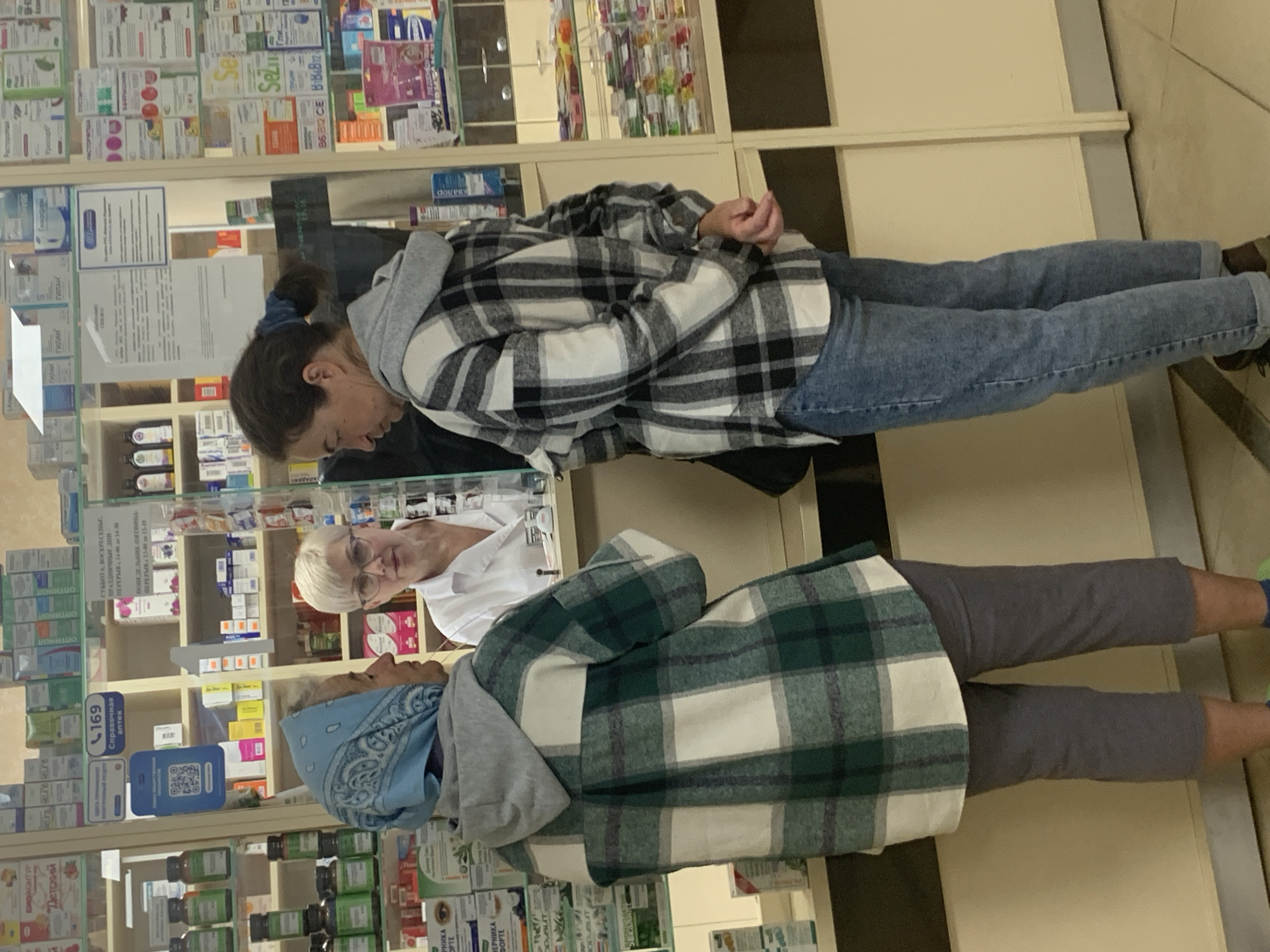 In a pharmacy in Kapyĺ, Belarus, two women stand at a counter interacting with a female pharmacist. The woman on the left, wearing a blue headscarf and a green and white plaid jacket over a grey hoodie, faces the pharmacist. The woman on the right, in a black and white plaid jacket and blue jeans, stands with her back to the viewer, also facing the pharmacist. The pharmacist, wearing a white coat and glasses, stands behind the counter. The pharmacy shelves behind them are stocked with various medications and health products. The environment appears to be a typical retail pharmacy setting.