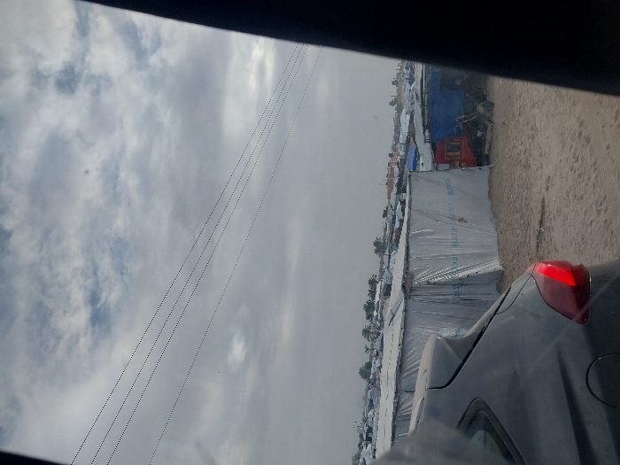 An overcast sky is visible with power lines extending across the view. Rows of white temporary shelters or structures stretch into the distance. A vehicle is partially visible in the foreground, with its taillight illuminated. A sandy area runs alongside the structures. The scene suggests a refugee camp or a temporary housing area, possibly photographed from within another vehicle.