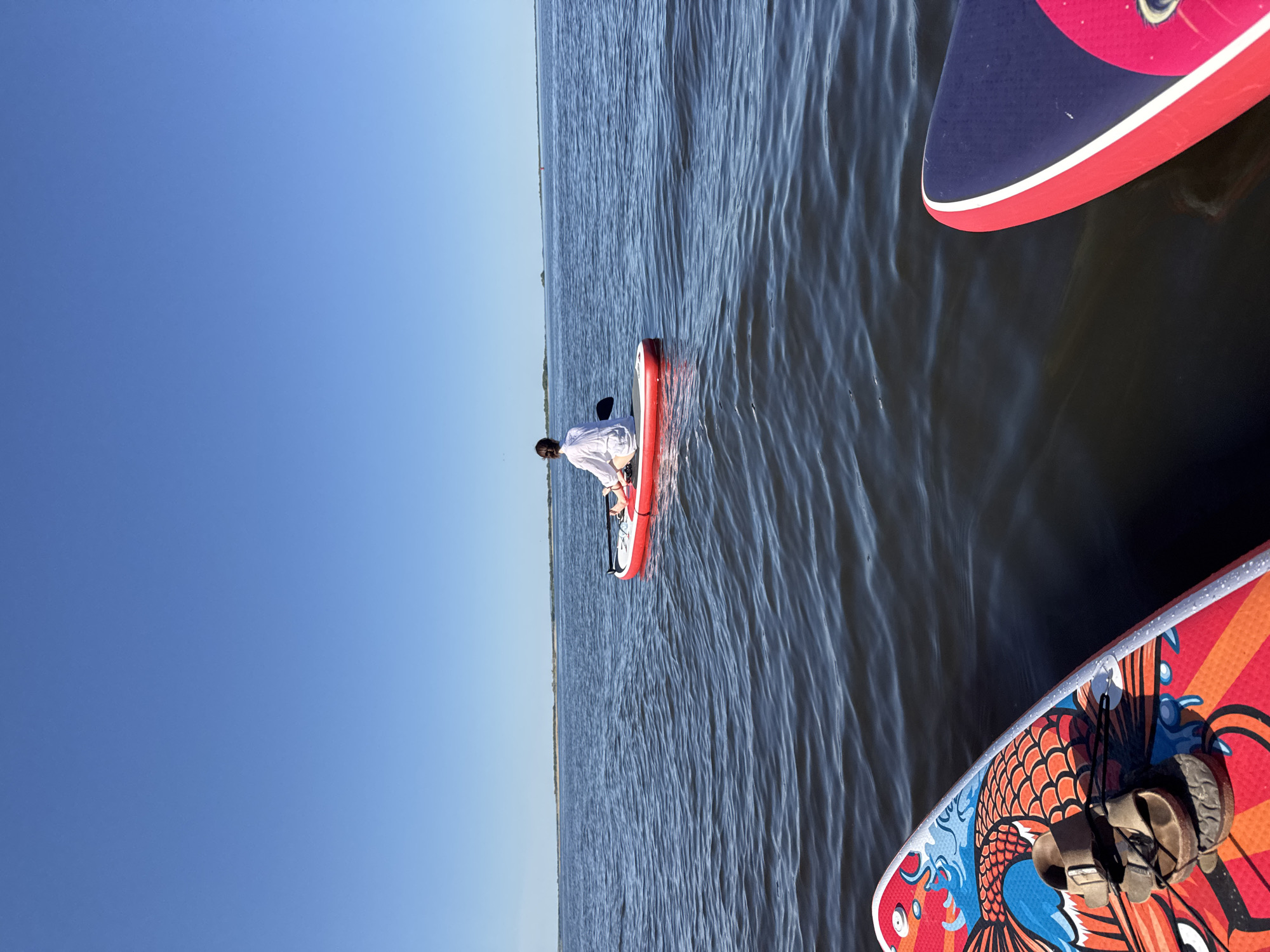 The image shows a body of water, possibly near Ust'-Kurdium, Russia, given the prompt context. A person sits on an orange paddleboard, facing away from the viewer. There are at least two other paddleboards nearby; one is predominantly red and blue, while the other has a colorful, possibly koi fish-themed, design, on which there is a pair of sandals. The sky is a clear blue. The water has visible ripples.