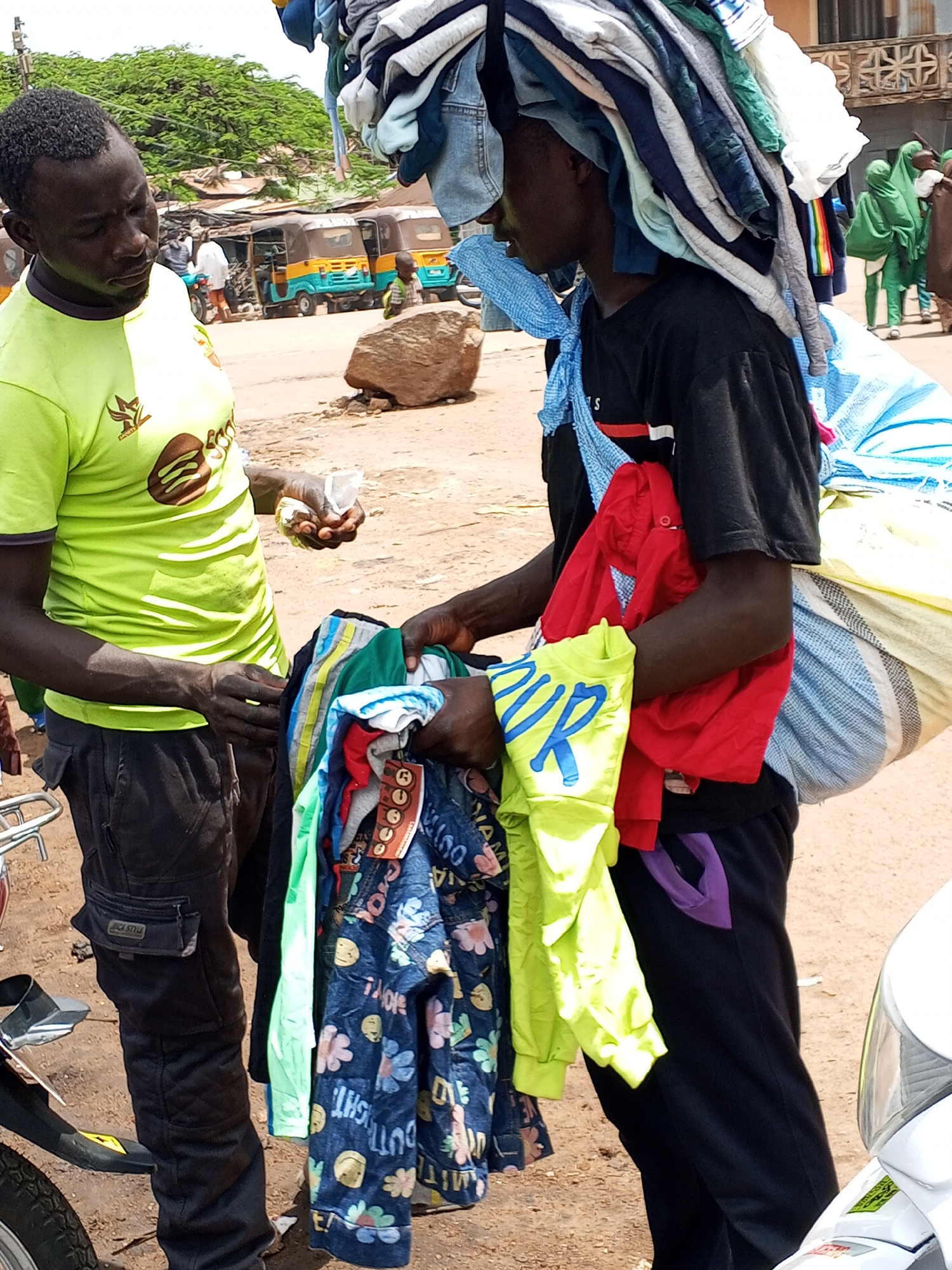 The image depicts two men in what appears to be a bustling marketplace in Jos, Nigeria. The man on the left wears a bright lime green t-shirt and dark pants. He is holding a small, wrapped item in his right hand and is looking down at it. The man on the right is carrying a large stack of clothes on his head, balanced by a blue cloth tied around his neck and shoulders. He is wearing a black t-shirt and dark pants, and a large, striped bag is slung over his right shoulder. He is holding a bundle of folded clothing in his hands.
The background shows several three-wheeled vehicles, commonly known as Keke Napeps, which are a popular mode of transport in Nigeria. There are also buildings and trees visible, suggesting an outdoor setting. The ground is unpaved, indicative of a typical marketplace. The overall scene suggests a busy commercial environment. The lighting indicates it is daytime, with what appears to be fair weather. The expressions of the men are neutral, focused on their tasks. The clothing on the man on the right has tags visible on some items, with text that is partially obscured but appears to include "OUT" and "GUT". The t-shirt of the man on the left has a logo with text that is difficult to discern but appears to be a stylized image.
