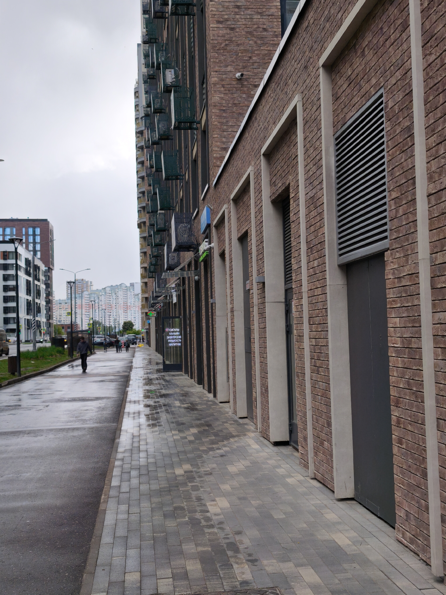 The image shows a street scene in Moscow, Russia on a cloudy day. The scene is dominated by modern brick buildings with prominent ventilation grilles. A wet sidewalk of gray brick pavers runs along the base of the buildings, reflecting the overcast sky. A narrow strip of wet asphalt road borders the sidewalk.
Several people are walking along the sidewalk. In the distance, high-rise buildings are visible, adding depth to the urban landscape. The image captures a typical urban environment, emphasizing the interplay of architectural design, pedestrian activity, and atmospheric conditions.