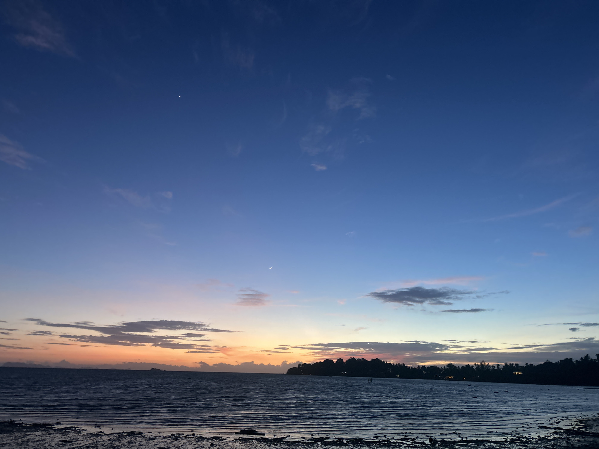 The image shows a serene coastal scene at dusk in Ko Pha-ngan District, Thailand. The sky transitions from deep blue at the top to soft oranges and pinks near the horizon, with scattered wispy clouds. A sliver of a crescent moon is visible high in the sky, along with a small, bright star. The calm sea reflects the colors of the sunset, with gentle ripples on its surface. In the distance, a dark silhouette of a treeline marks the shore, with a few faint lights suggesting human habitation. The foreground shows a sandy, wet beach with small stones and debris. Two distant figures, barely discernible, are standing in the shallow water near the shore. The overall atmosphere is tranquil and peaceful, indicative of the end of the day.