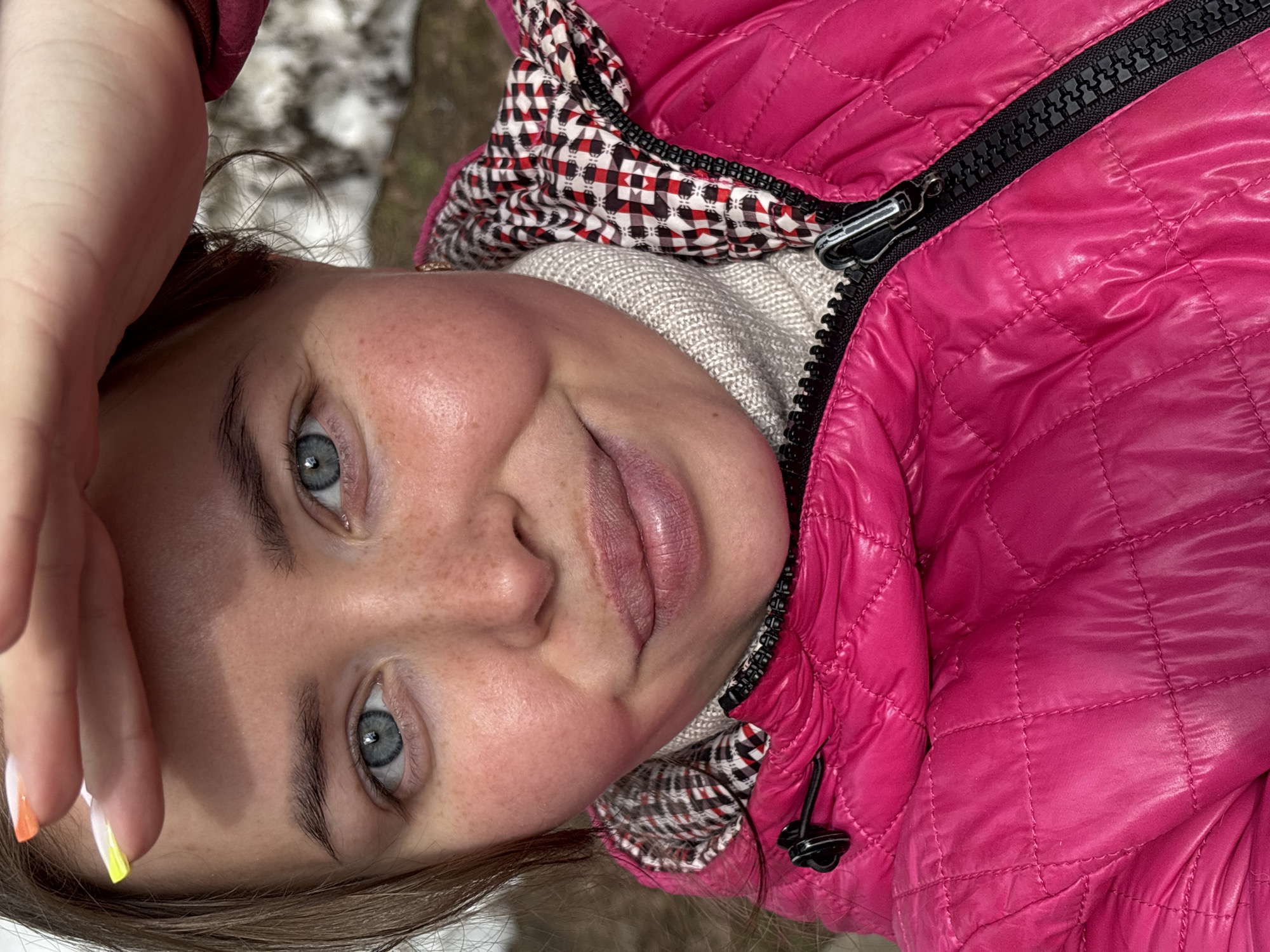 A close-up selfie of a woman wearing a bright pink quilted jacket with a black zipper and a beige turtleneck sweater. The jacket hood has a black, white, and red checkered pattern lining. Her blue eyes are visible, and she has freckles. Her hand is raised to her forehead. The location is not discernible from the image. Русский Вожой, Russia is not confirmed.