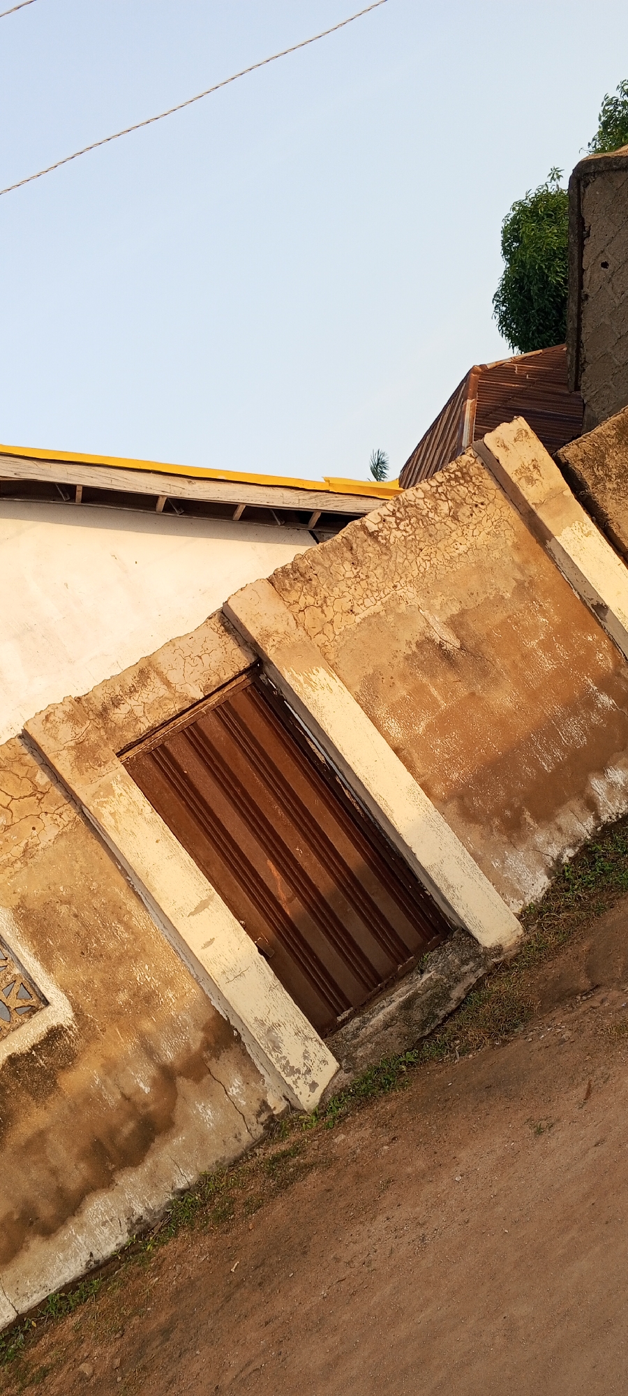 A low-angle, Dutch-tilted outdoor shot depicts the exterior of a weathered building under a clear, pale blue sky. Two thin, parallel electrical wires stretch across the upper portion of the frame, angled slightly from left to right.
In the mid-ground, a section of a building features a brown, ribbed metal door set within a rough, concrete-rendered wall. The wall exhibits a textured, cracked surface with areas of discoloration and peeling paint, primarily in shades of tan and off-white. Two concrete pillars, one on either side of the door, are integrated into the wall; they are painted in a faded cream color and show signs of wear and tear. To the left of the door, a small, decorative lattice-like opening is visible in the wall.
Above the concrete structure, a portion of a white wall with a bright yellow edge is visible. Further to the right, a corrugated metal roof, appearing rusty and aged, protrudes from behind another building. A small, green tree with dense foliage is situated behind and to the right of the corrugated roof, peeking over the edge of a more substantial, rough-textured stone or concrete building.
The foreground is dominated by a sloping dirt path, which appears to be made of reddish-brown soil. Patches of sparse green grass and weeds are growing along the edge where the path meets the building's concrete base. The lighting suggests it is late afternoon or early morning, casting long shadows and highlighting the textures of the surfaces. The scene appears to be located in Jalingo, Nigeria, given the architectural style and environmental elements. No people are visible in the image.