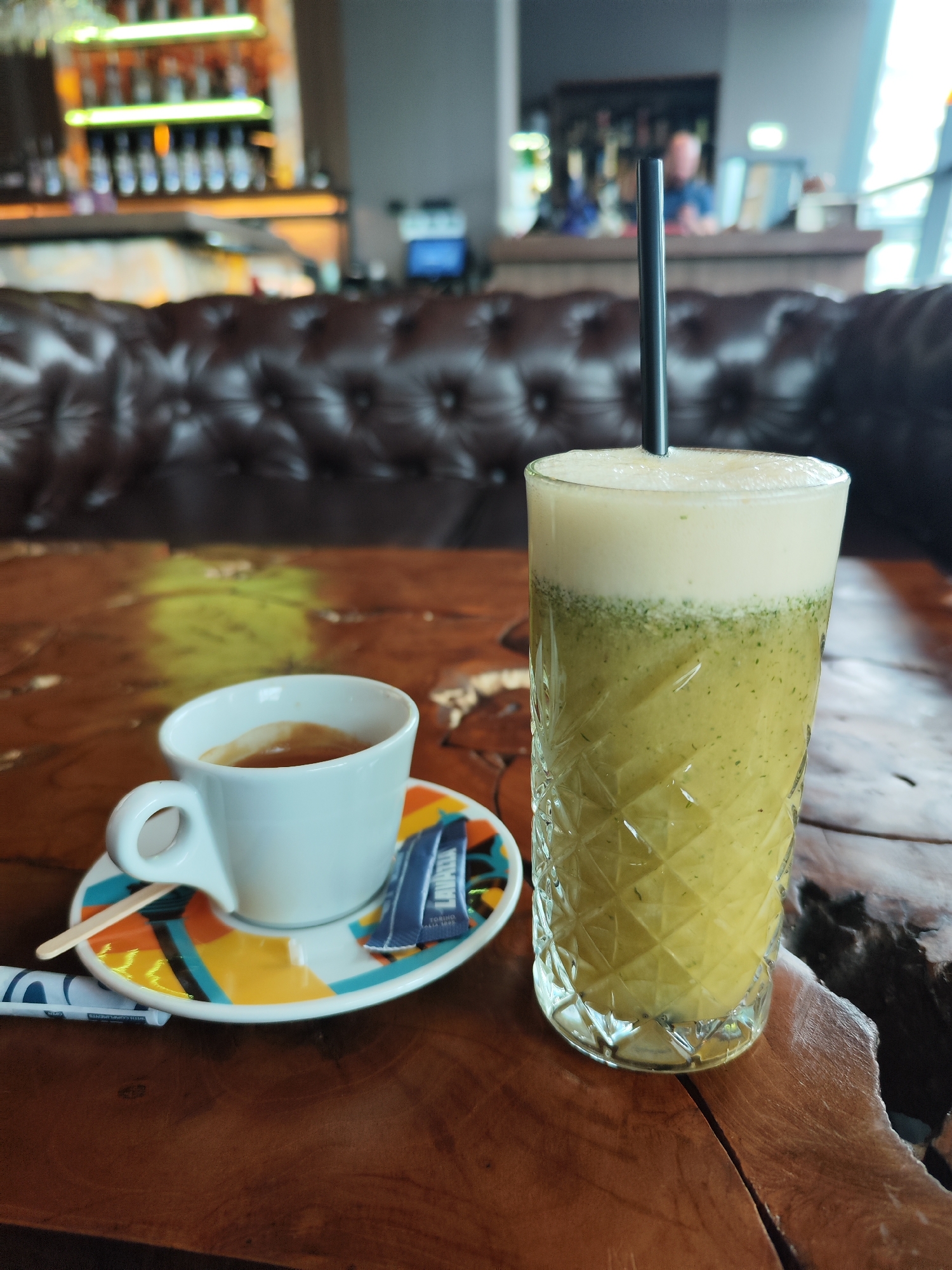 A close-up shot shows a cup of espresso coffee and a tall glass of a green-and-white layered drink with a black straw on a wooden table. The coffee sits on a saucer with a colorful geometric design, and there is a packet of Lavazza sugar. In the background, a dark brown leather sofa and a bar area with bottles are visible, suggesting the location is a café or bar in Burgas, Bulgaria. There are no people clearly visible in the foreground, but a blurred figure of a person can be seen behind the bar.