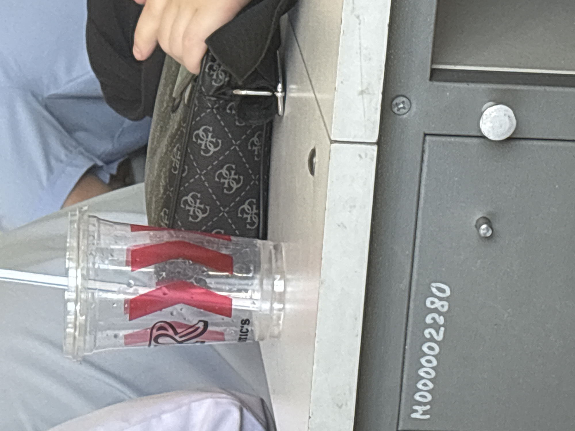 The image, rotated clockwise, captures a close-up indoor scene focusing on a person's hand, a handbag, and a drink cup positioned next to a piece of equipment or furniture.
A person, visible from the mid-torso down, is wearing light blue clothing, possibly scrubs or casual pants, and has light-toned skin on their visible hand and arm. Their hand is resting on or holding a black handbag adorned with a repeated "GUESS" logo pattern.
Next to the handbag is a clear plastic cup containing a beverage and a straw. The cup features red graphic elements and partial text, including a stylized "R" and what appears to be "IC'S". The cup is resting on a light gray or white surface, possibly a counter or table.
To the right of the bag and cup is a dark gray or black surface, likely a piece of industrial equipment, a locker, or a mailbox, secured with several screws and featuring a prominent white, circular knob or fastener. On this dark surface, a series of numbers is written in white: "M000002280". The overall scene suggests a moment of rest or waiting in a public or semi-public indoor setting, possibly an office, medical facility, or transport hub.