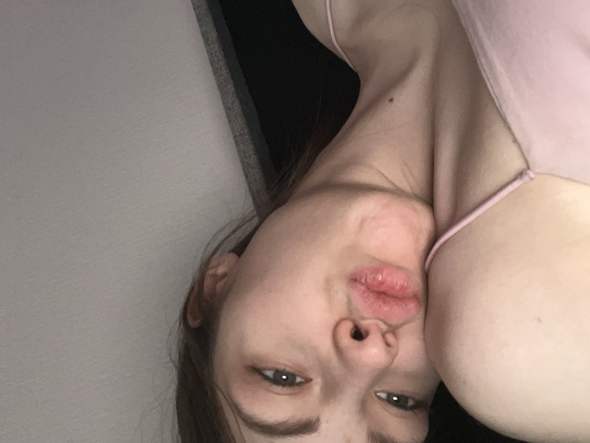 A close-up, upside-down photograph features a person with fair skin, light brown hair, and green eyes, who appears to be female. The subject is wearing a light pink top with thin straps. A portion of the person's chest is visible. The subject's lips are prominently displayed and appear to be irritated or chapped. The background is indistinct, showing a light-colored wall and a darker area near the top. The photograph provides no contextual information suggesting a location in Saint Petersburg, Russia or any other identifiable place.
