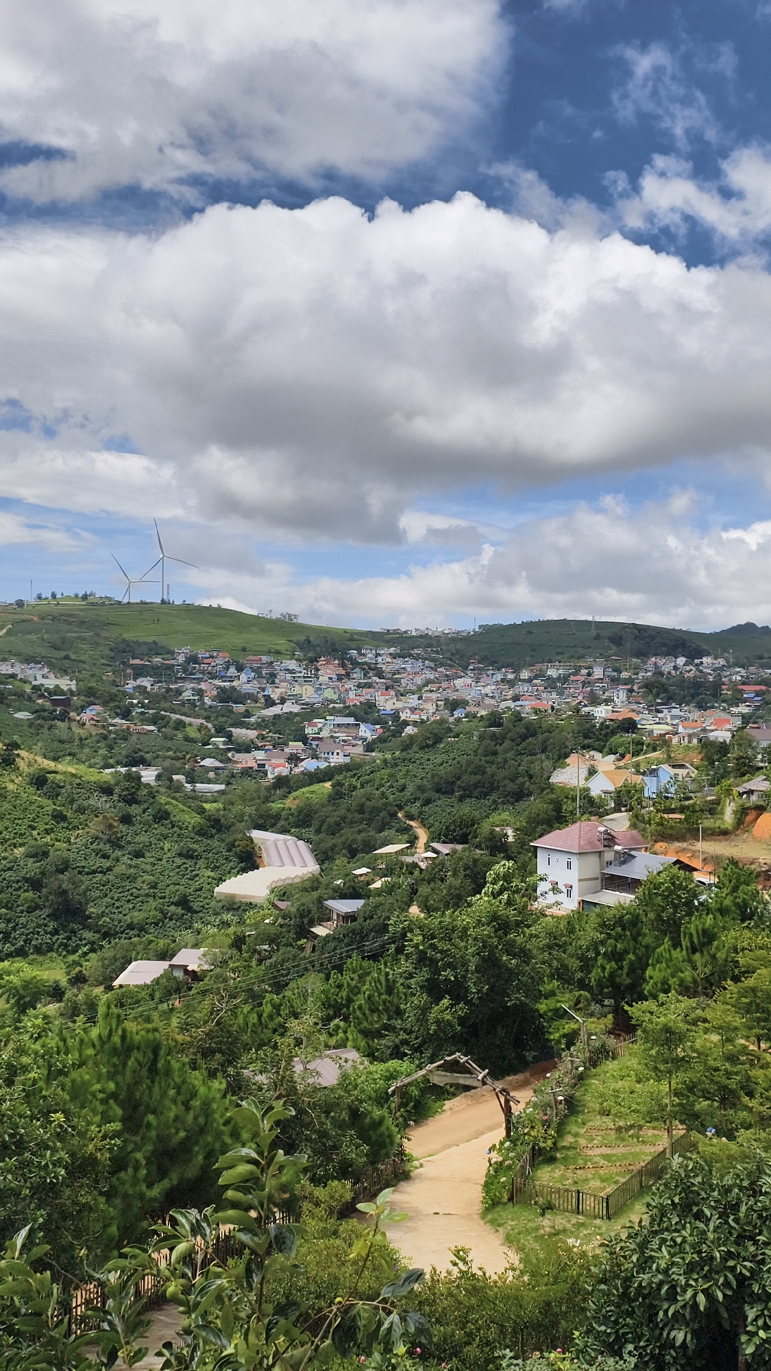 The image captures a panoramic view of a hilly landscape in Đa Thọ, Vietnam, under a partly cloudy sky. In the foreground, lush green trees and foliage dominate, with a dirt path winding through the vegetation, leading to a wooden archway. Beyond the immediate foreground, the landscape slopes downwards, revealing a densely populated town nestled in the valley. The town is characterized by a collection of buildings with colorful roofs, indicative of residential housing. Further in the distance, on the crest of a hill, two wind turbines stand as prominent features against the sky, suggesting a source of renewable energy. The overall scene depicts a harmonious blend of natural scenery and human habitation, with the town integrated into the rolling hills.