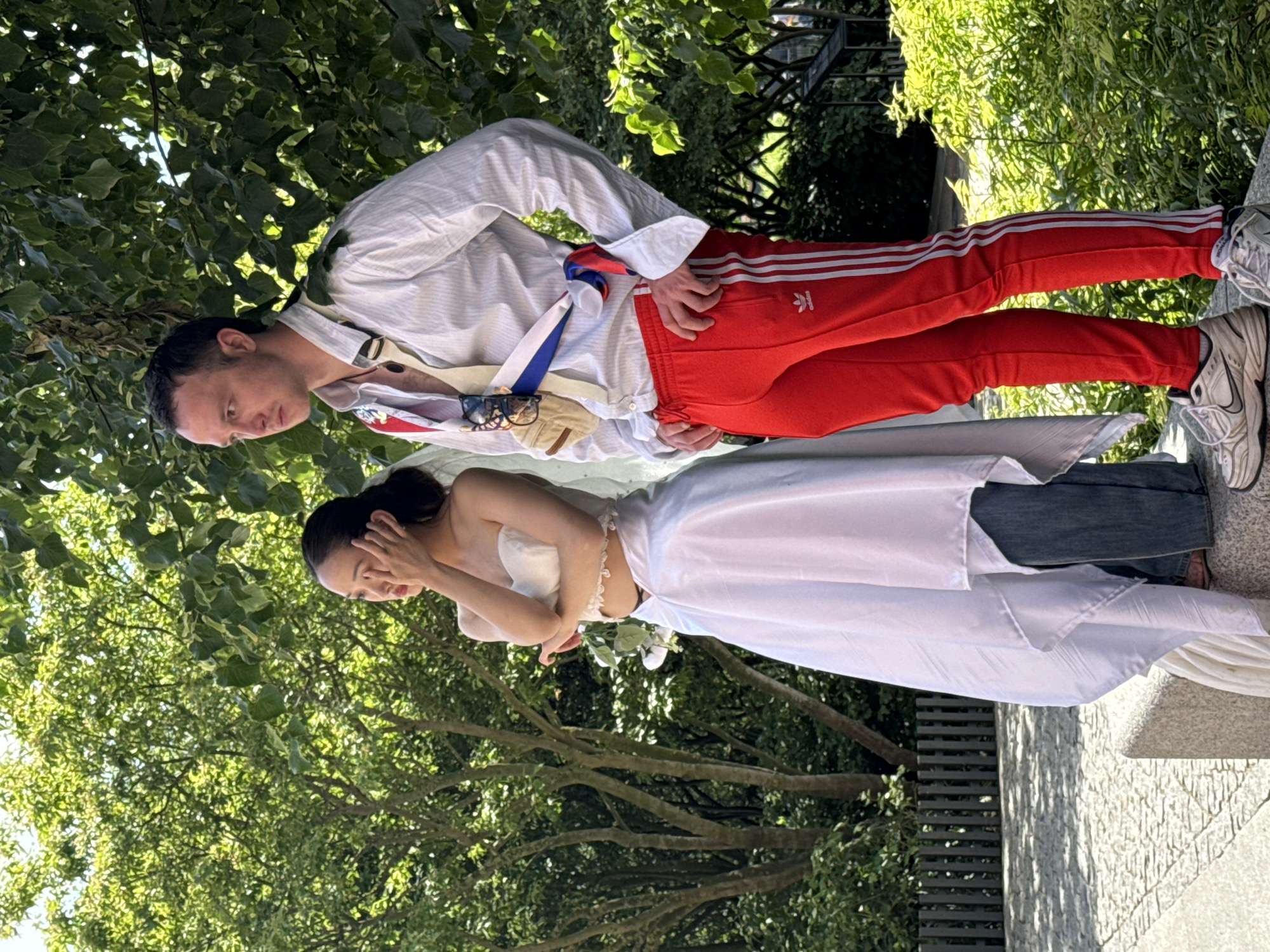 Two people stand outdoors. The person on the left appears to be a woman wearing a white strapless top and a white skirt over blue jeans. Her right hand is touching her face. The person on the right appears to be a man wearing a white pinstripe shirt, red track pants with white stripes, and white sneakers. A sash with blue and white stripes is draped across his chest, and a beige pouch is attached to his shoulder. Both individuals are looking to their left. They are positioned on a stone surface, with trees and foliage in the background. A metal fence is visible to the left of the frame. The scene is bathed in daylight. There is no indication of the location being Saint Petersburg, Russia.