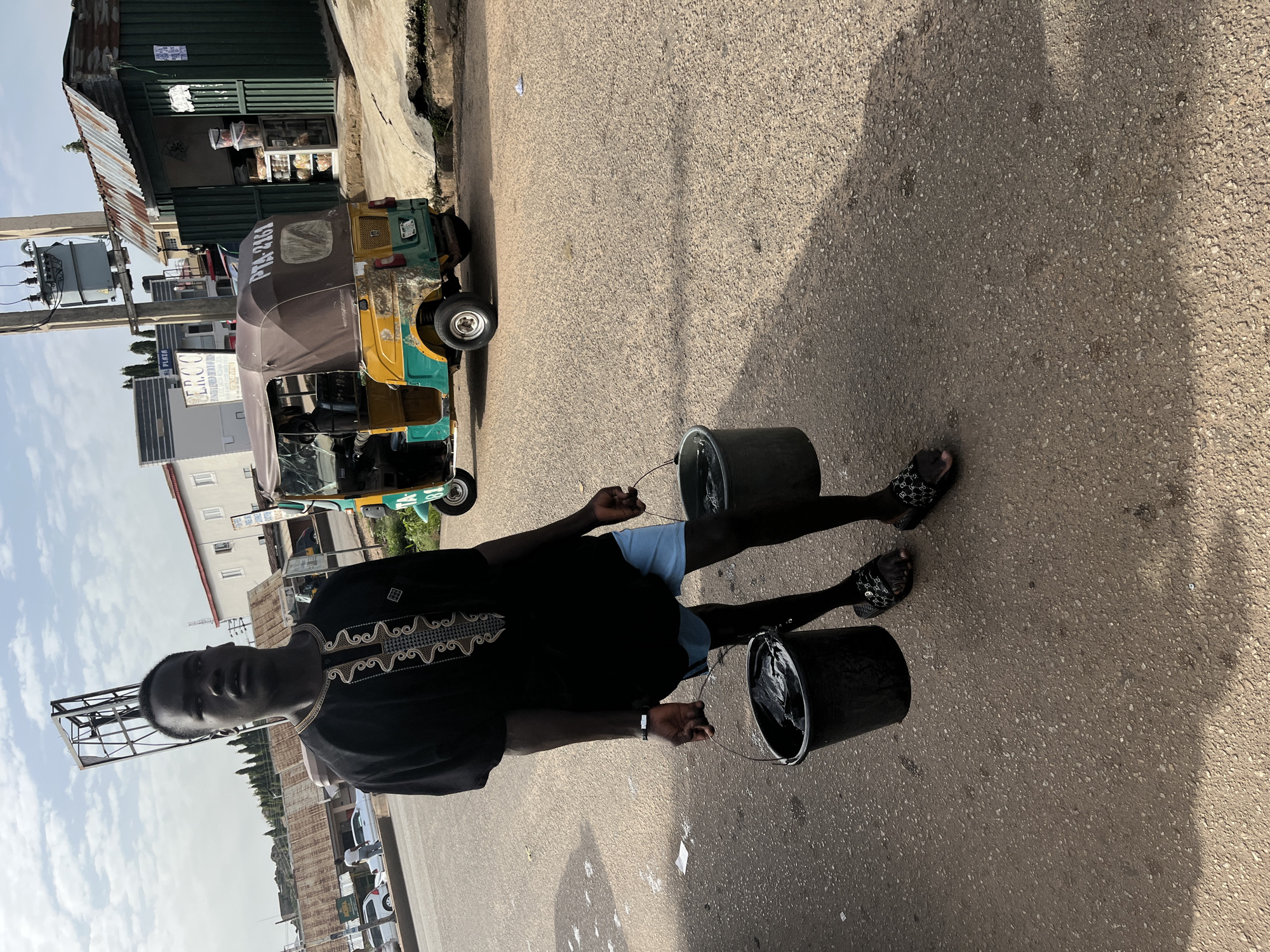 A young Black male stands on an asphalt road in Jos, Nigeria, carrying two dark buckets filled with water. He is wearing a black traditional African garment with white and gold embroidery on the chest and light blue shorts. The buckets are positioned on either side of his legs. In the background, a colorful auto-rickshaw with the license plate "PTA 2161" is parked. Further back, there are several buildings, including one with a corrugated metal roof that appears to be a shop. A utility pole with a transformer stands to the left of the shop. The sky is partly cloudy.