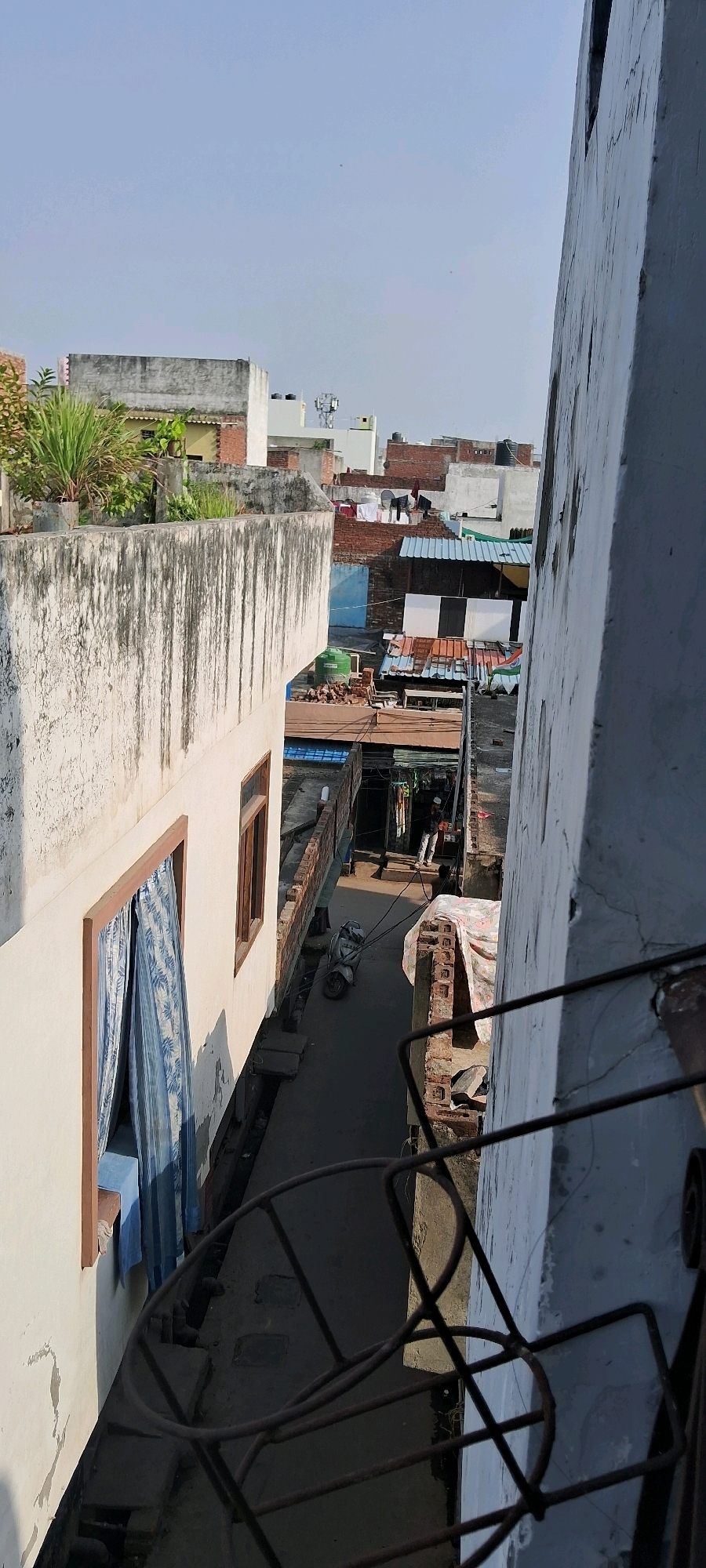 This vertical, eye-level, outdoor photograph captures a narrow alleyway between buildings in a densely populated urban area, likely in Meerut, India. The scene is characterized by a mix of residential and commercial structures, with a clear view of the sky.
In the foreground, on the left, is the side of a light-colored building with a textured, weathered facade. A window with blue patterned curtains is visible. To the right of this building, a dark, metal structure, possibly a railing or part of a discarded object, occupies the lower portion of the frame.
The alleyway itself is paved and appears to be a service or pedestrian passage. A silver scooter is parked on the right side of the alley. Further down the alley, the scene becomes more cluttered with various structures, including corrugated metal roofs, brickwork, and what appear to be makeshift shelters. Laundry is hung out to dry on lines between buildings.
In the background, more buildings of varying heights and construction materials are visible. Some have water tanks on their roofs. A person wearing a white shirt and dark pants, possibly a man, is walking away from the camera in the alley. Another person is partially visible further down the alley.
The overall impression is one of a busy, lived-in urban environment with a strong sense of community and everyday life unfolding. The lighting suggests it is daytime, with the sun casting shadows. The clear, pale blue sky indicates good weather.