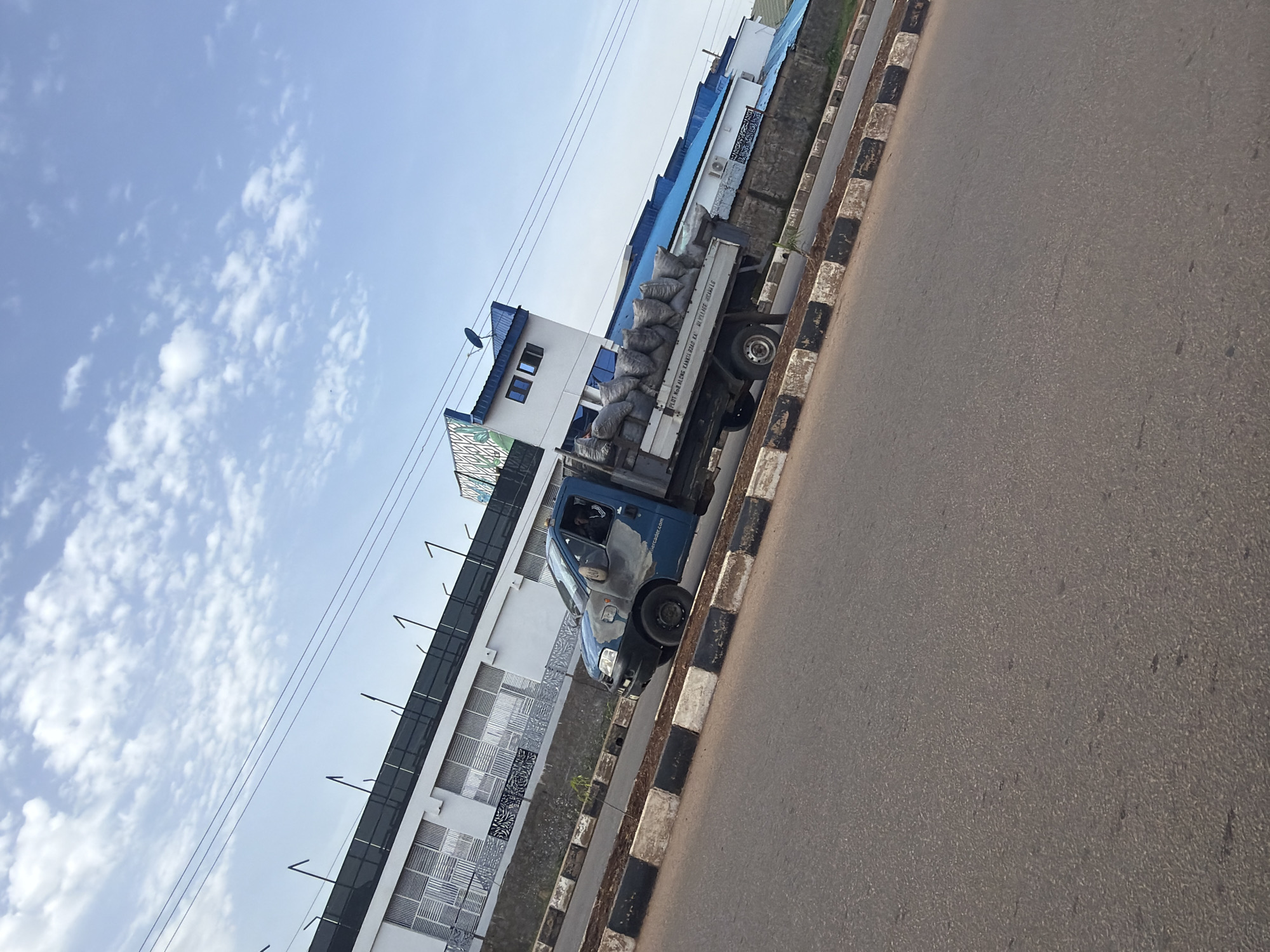 A blue pickup truck is parked on the side of a road, its bed filled with several large sacks, likely containing goods. The truck has a driver inside. The vehicle is positioned alongside a building with a blue roof and white walls, featuring decorative patterns on some sections. The road is paved, with a black and white striped curb separating it from the building's property. Overhead, a clear blue sky is dotted with scattered white clouds. The scene appears to be a sunny day, likely during daylight hours. A visible inscription on the side of the truck reads "FLOT NG8 ALONG KANTI ROAD NIKELAGE NIGERIA". The image was taken in Gura Top, Nigeria.