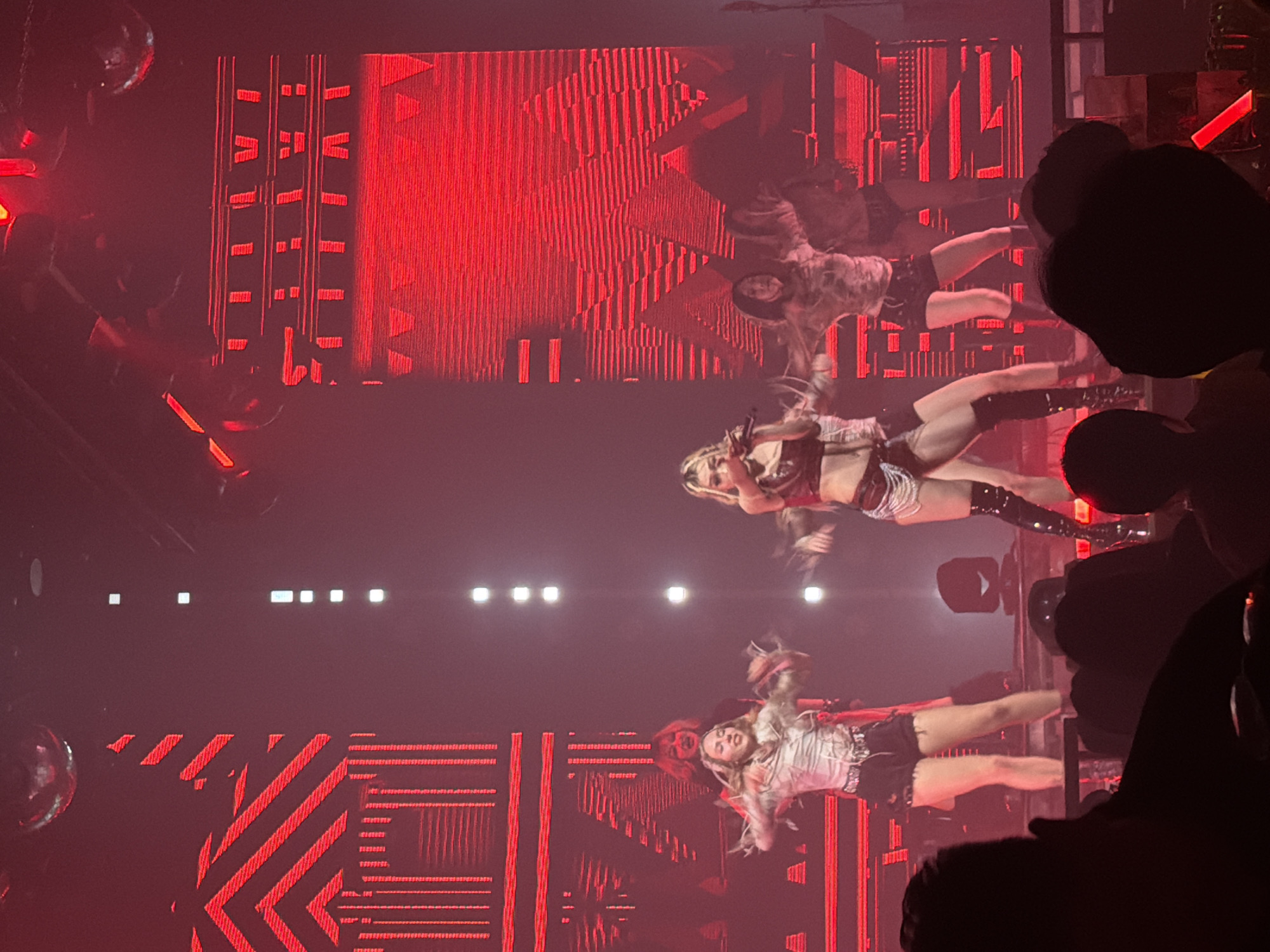 The image captures a live performance on a stage within a venue in Jiangbeicheng Subdistrict, China, characterized by dramatic red lighting. Approximately seven individuals are visible on stage, actively performing. In the center, a blonde-haired individual, likely the lead performer, is holding a microphone to their mouth, dressed in a dark, possibly leather, crop top, dark shorts adorned with silver chains, and knee-high dark boots. This individual is positioned slightly forward with their right leg bent. To the left, another female performer with light hair wears a white corset-style top, dark shorts, and is captured mid-movement with arms raised. Behind the central figure and to her left, a person with red hair is partially obscured, wearing dark clothing. Other backup dancers are visible across the stage, some in patterned tops and dark shorts, performing synchronized movements.
The stage background consists of large LED screens illuminated with vibrant red geometric patterns, including vertical lines, horizontal bars, and chevron shapes, creating a dynamic visual effect. A vertical strip of bright white rectangular lights extends upwards from the stage floor in the central-right portion of the background. A disco ball is partially visible in the upper-left corner of the frame. The foreground shows the silhouetted heads and shoulders of an audience, indicating a close perspective from the crowd. One audience member on the far bottom right holds a glowing orange/red light stick. The overall environment is dark except for the stage lighting.