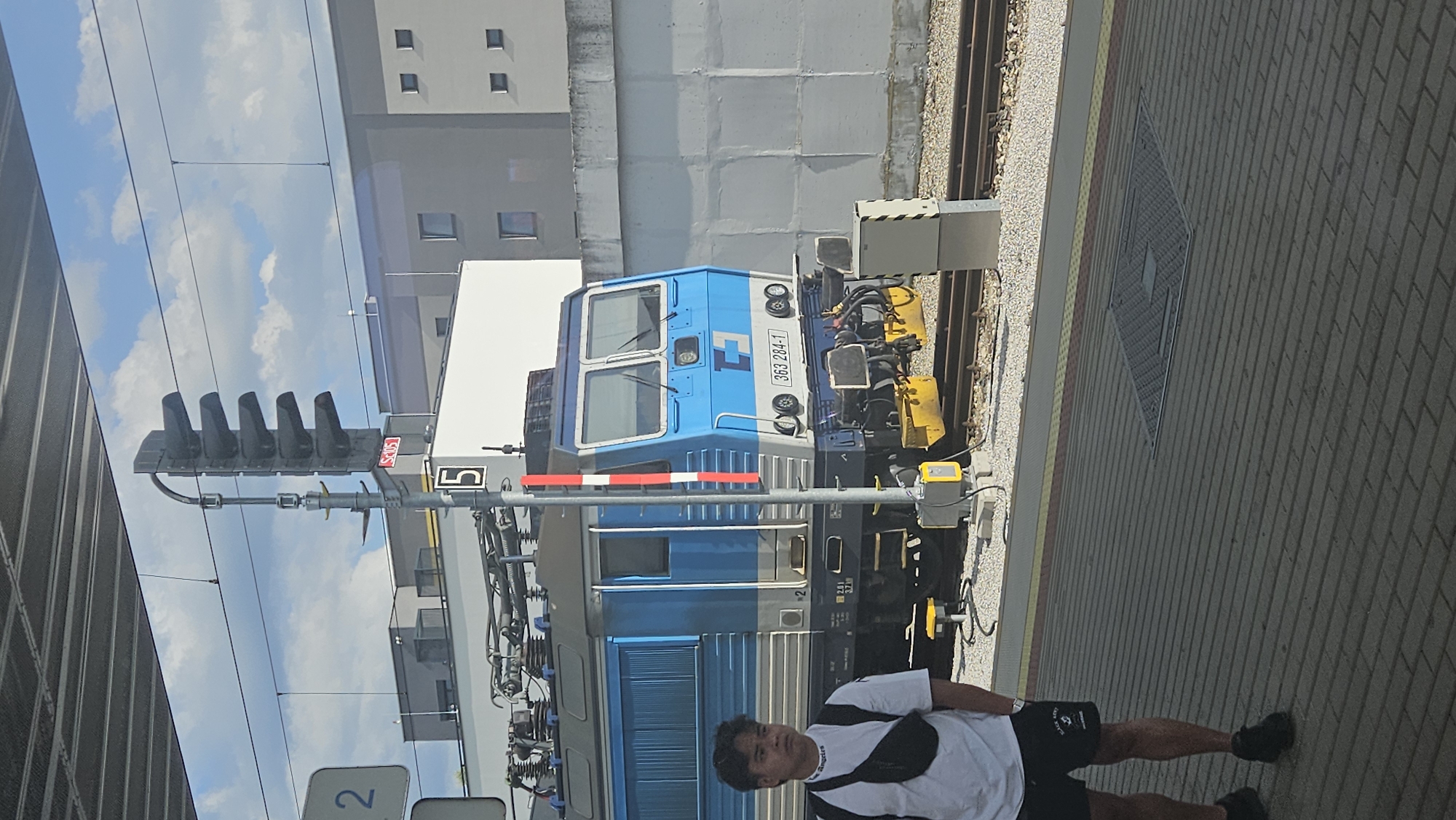 A young man stands on a train platform in Trnava, Slovakia, to the left of a blue and white electric locomotive. He is wearing a white t-shirt, black shorts, and black sneakers. A black fanny pack is worn across his chest. The train is numbered "363 284-1". Behind the train is a grey concrete wall and modern buildings. A train signal with multiple lights stands to the left of the locomotive. The platform is paved with grey bricks, and there are train tracks in front of the locomotive. The sky is blue with some clouds.