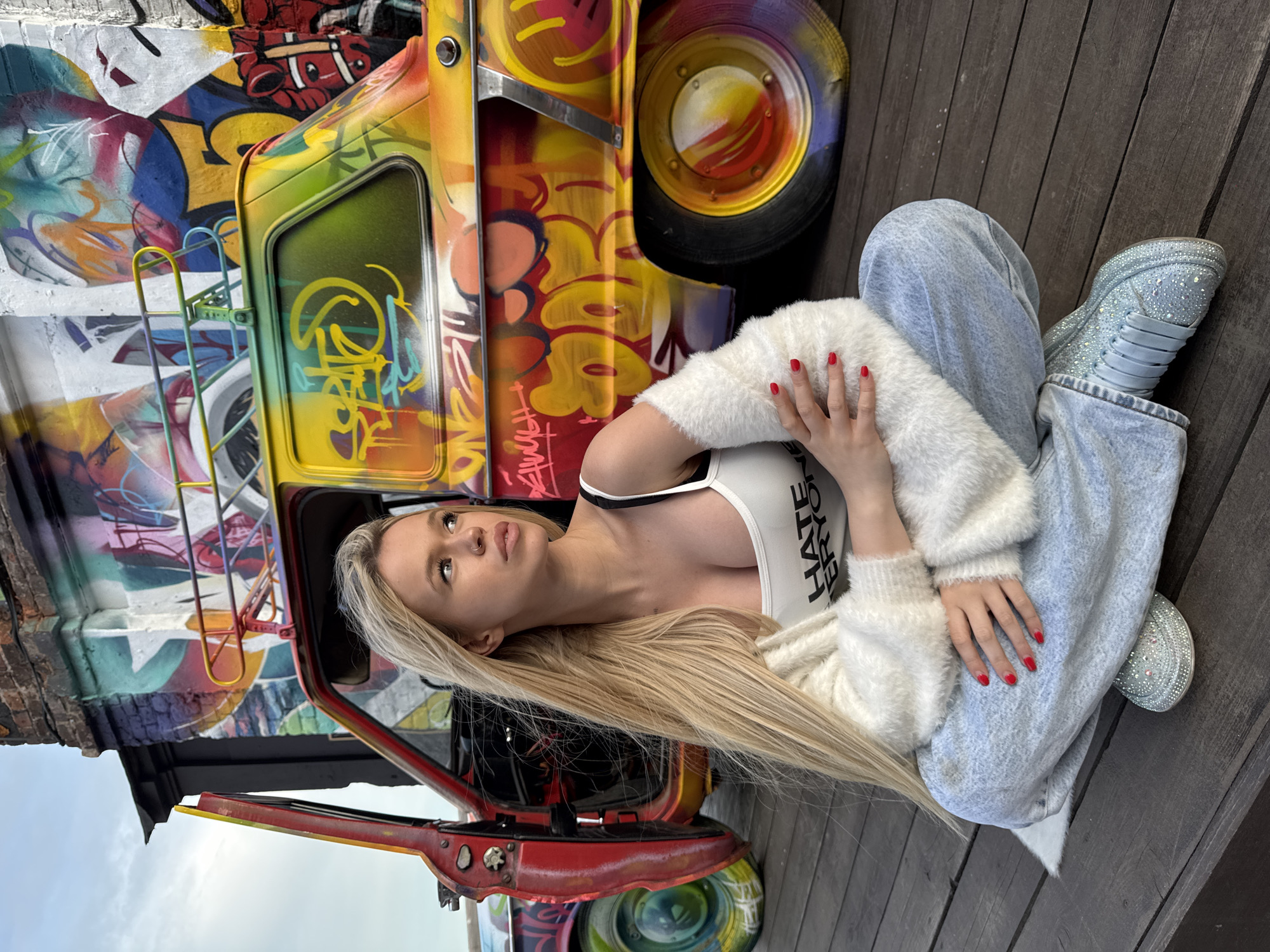 A young woman with long blonde hair sits cross-legged on a wooden deck in front of a brightly painted vintage car covered in graffiti. The car, a retro Soviet-era model, is adorned with vibrant, colorful spray paint art. The woman wears a white tank top with the text "HATE EVERYONE" printed on it, a fluffy white cardigan, and light blue denim pants. Her toenails are painted red, and she wears sparkly sneakers. The background features more graffiti on a brick structure and a cloudy sky. The scene suggests an urban, artistic setting, possibly an outdoor exhibition or a street art location in Saint Petersburg, Russia.