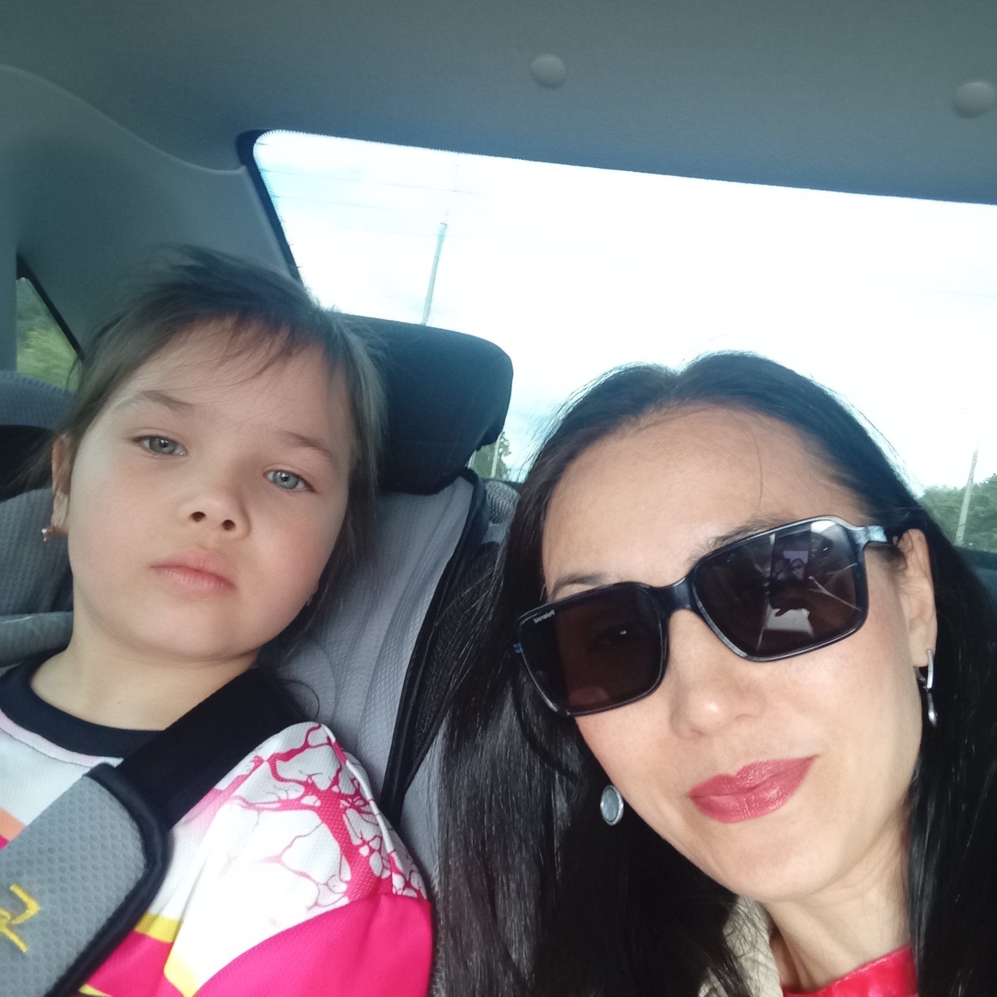 The image depicts an adult female and a young female child inside a vehicle in the city of Pervomaysky, Russia. The adult female, positioned in the foreground and appearing to be taking a selfie, has dark, long hair, is wearing dark sunglasses, red lipstick, and visible earrings (a silver hoop and a round blue/grey stud). Her reflection in the sunglasses shows her holding a smartphone. She is wearing a red collar or neckline over a lighter-colored garment. The young female child is seated in the background, secured by a car seatbelt with a grey padded protector. She has light brown hair, light-colored eyes, and a neutral expression, looking directly at the camera. She is wearing a colorful shirt, predominantly white with pink/red tree-like patterns and yellow accents, over a black shirt. The car's light-colored headliner is visible above them, and through the rear window, an overcast sky, distant greenery, and a thin pole-like structure are discernible. The interior elements like car seats and headrests, in shades of grey and dark blue/black, are also visible.
