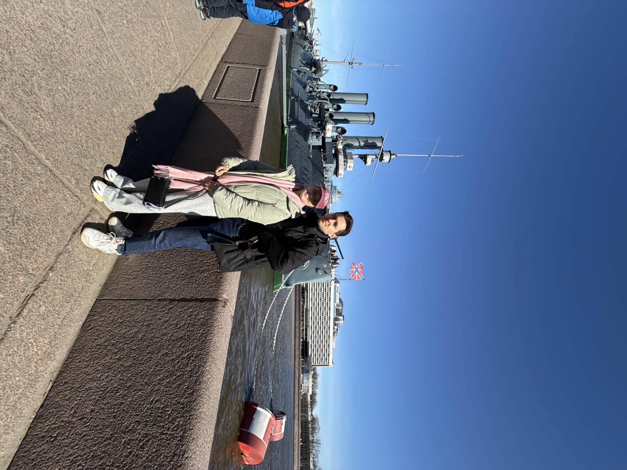 The image shows a couple seated on a granite embankment along the Neva River in Saint Petersburg, Russia. The woman wears a light green jacket and a pink scarf and hat. The man is in a dark jacket. They are positioned in front of the Cruiser Aurora, a historic Russian warship. In the background are the blue sky, a modern building, and other unidentified people standing near the cruiser. In the river, a red and white buoy is visible.