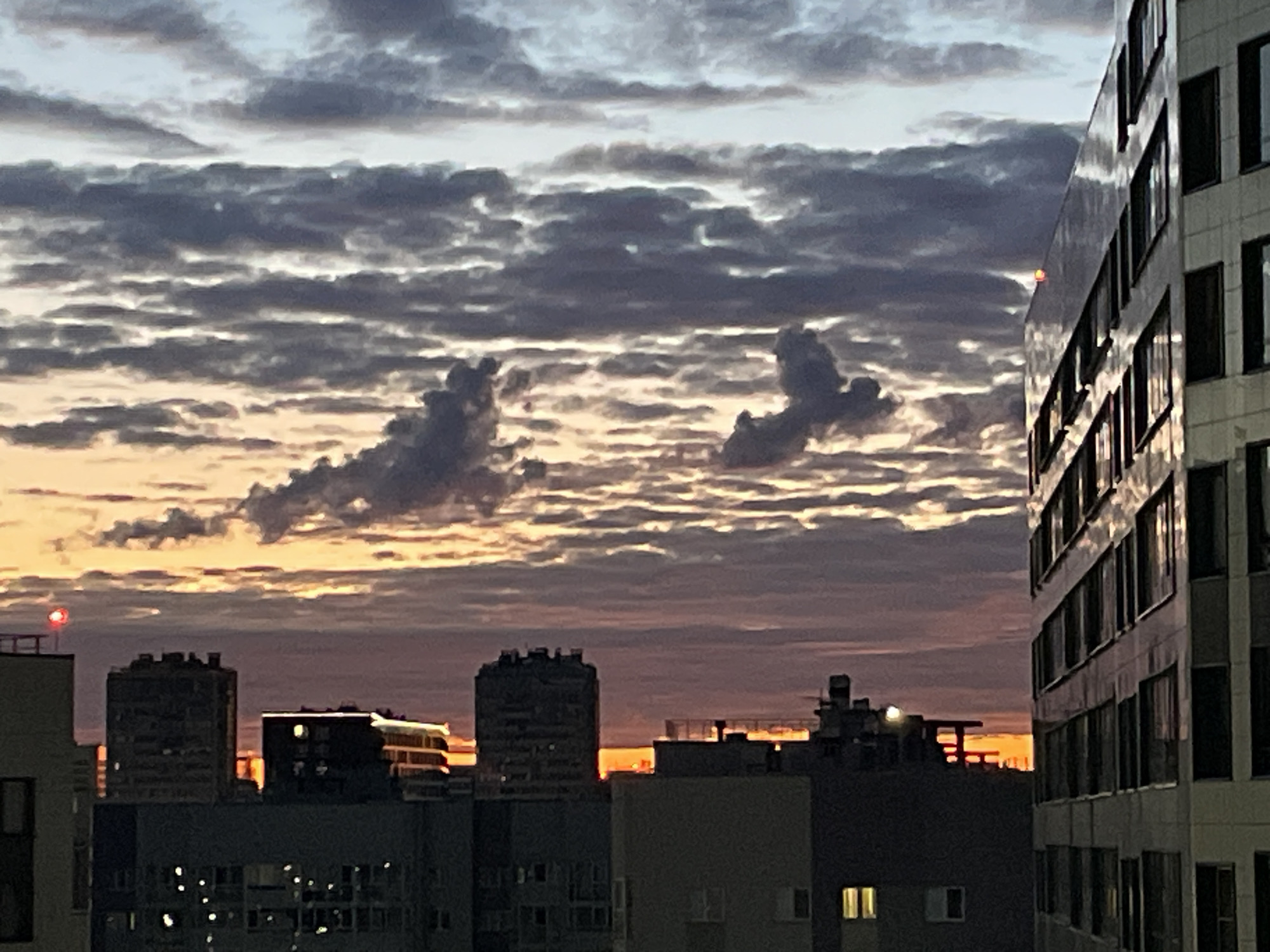 This is a photograph of a city skyline at dusk. The sky is filled with dramatic clouds, illuminated by the setting sun, casting warm orange and yellow hues. Two distinct cloud formations in the center of the frame resemble abstract figures. In the foreground, a modern, multi-story building with numerous windows dominates the right side of the image. To the left and center, other buildings of varying heights are visible, some with illuminated windows suggesting occupancy. The overall atmosphere is serene and captures the transition from day to night in an urban environment. The time of day is clearly dusk, indicated by the low sun angle and the colors in the sky. The weather appears to be partly cloudy, with the clouds creating interesting textures and light patterns. There is a red flashing light visible on top of a building on the left. No text is visible in the image.