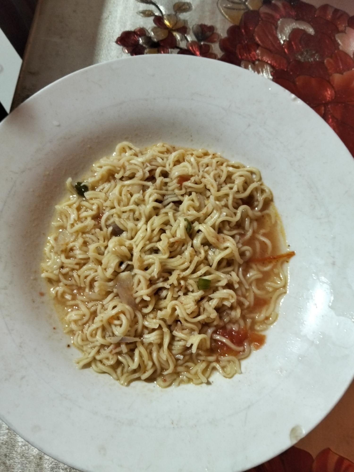 A close-up, top-down shot shows a white bowl filled with instant noodles and broth. The noodles are golden yellow and coiled within the shallow bowl, which is resting on a patterned tablecloth. The broth appears to be a light brown color and contains visible pieces of what look like tomato and possibly onion or other vegetables. A few strands of green onion are also visible. The lighting suggests it could be an indoor setting, possibly a home or restaurant. The background shows a decorative item with red and gold petals. No people are visible in the image. The image focuses solely on the food, suggesting it is a culinary or food-related shot. There is no visible text in the image. The setting appears to be in Payagaboh, Indonesia.