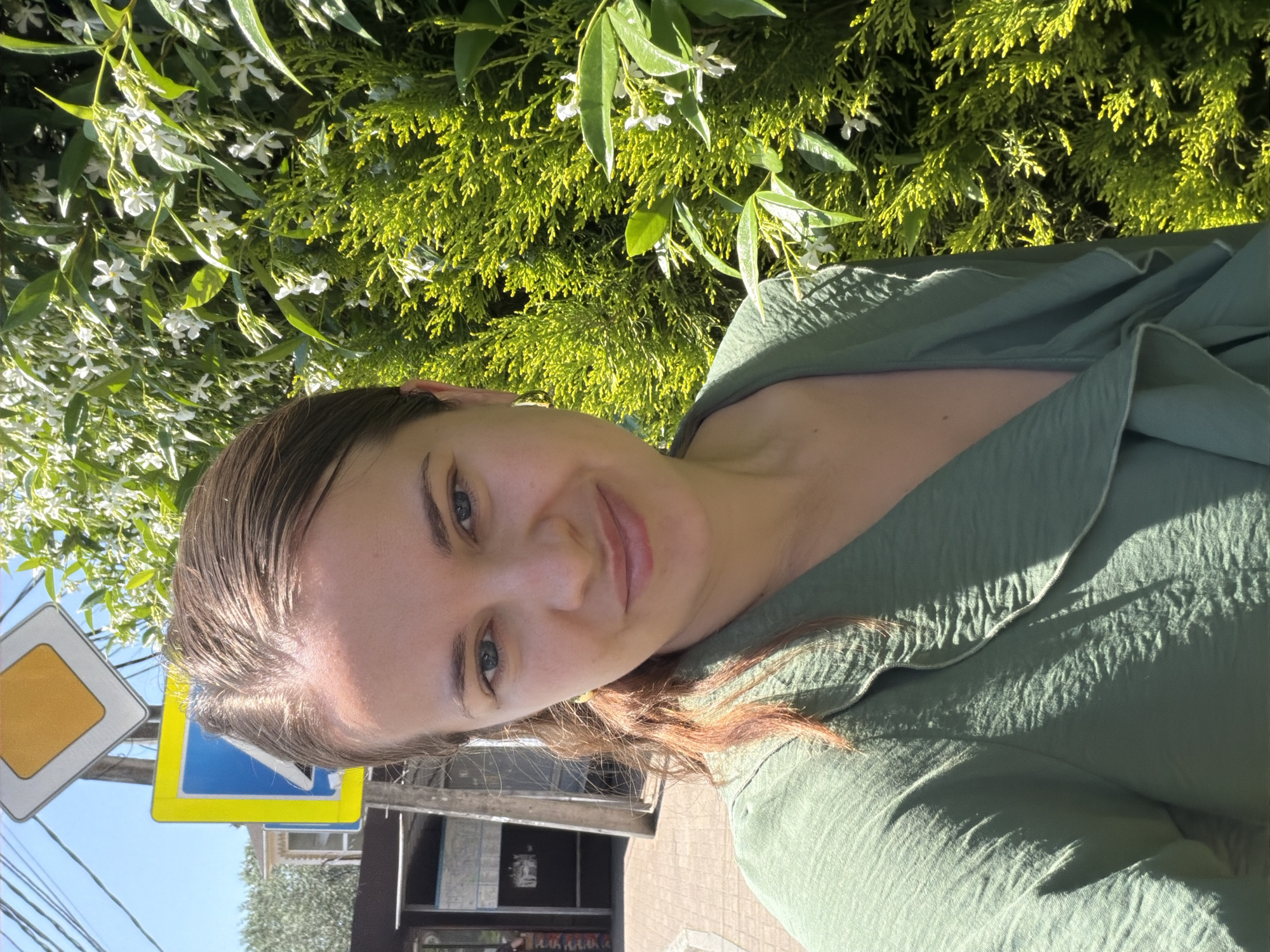 A young woman with medium brown hair pulled back into a ponytail smiles at the camera. She is wearing a green collared blouse and a light-colored earring. Behind her, on the left, is a city street with a sidewalk, a building with a sign, and a traffic sign pole. On the right, a lush green bush with white flowers is visible. The sky is clear and blue, suggesting a sunny day. The image appears to be a selfie taken outdoors.