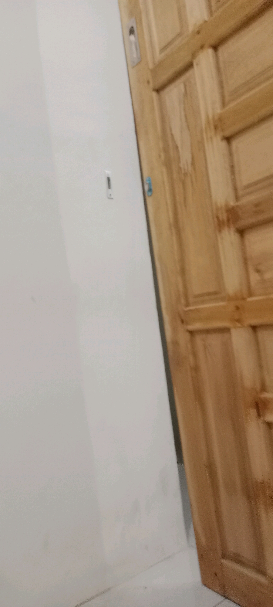 A wooden door is partially visible on the right side of the image, meeting a white wall on the left. The door appears to be made of lighter-colored wood with distinct grain patterns and paneling. A small, rectangular metallic object is embedded near the top of the door. On the white wall, a similar metallic object is mounted horizontally, with a smaller vertical indicator above it. The lighting suggests an indoor environment. The scene appears to be a hallway or entryway.
