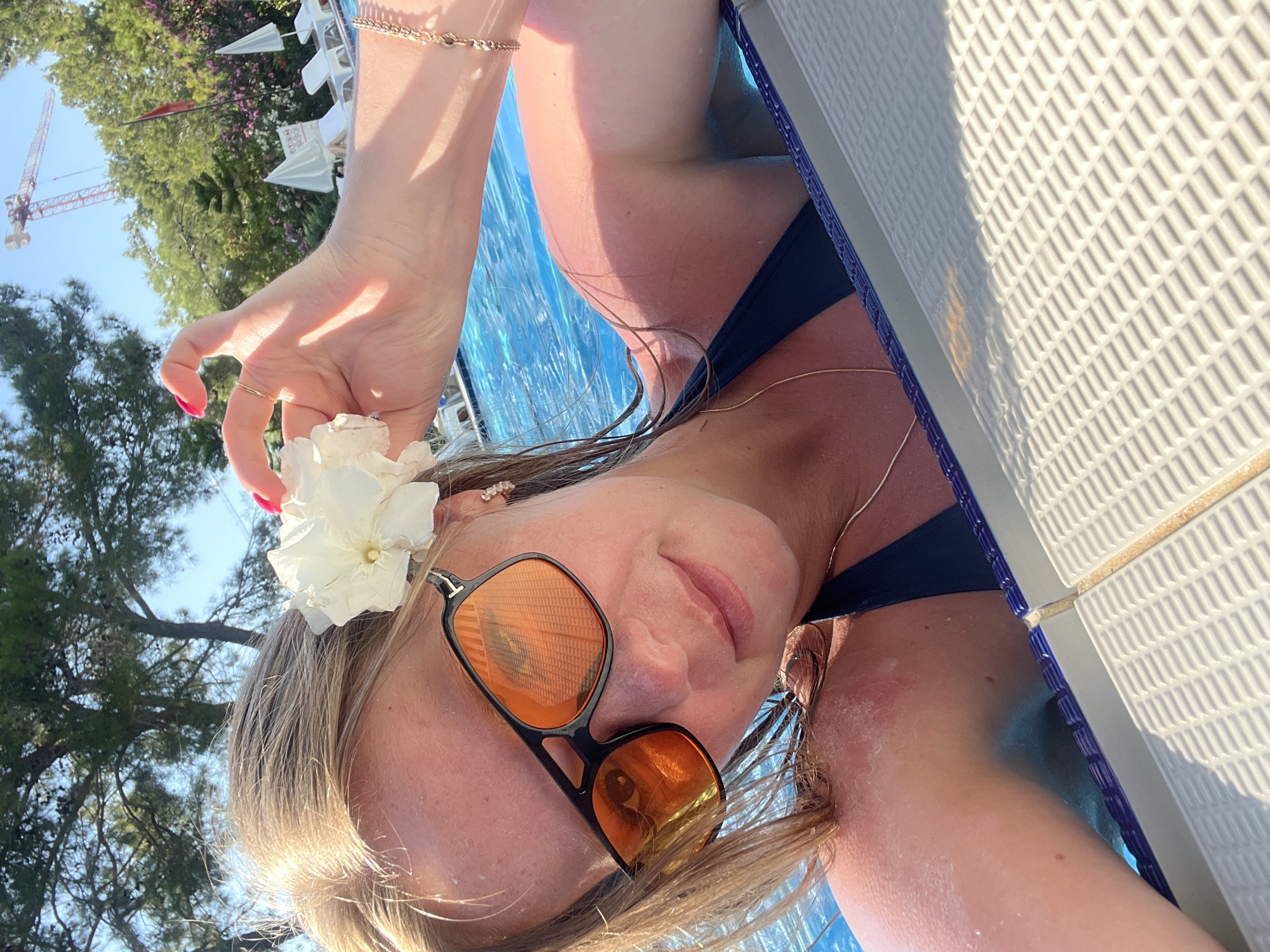 A young woman with blonde hair and sunglasses is taking a selfie in a swimming pool. She is wearing a dark swimsuit and has a white flower in her hair. The background shows trees, lounge chairs, and a construction crane. The image appears to be taken in Alanya, Turkey, as indicated by the presence of a construction crane, which is a common sight in rapidly developing tourist areas. The woman's skin shows signs of sunburn on her shoulders, suggesting a sunny day. The water in the pool is a vibrant blue, and the sunlight reflects off its surface, creating a shimmering effect.