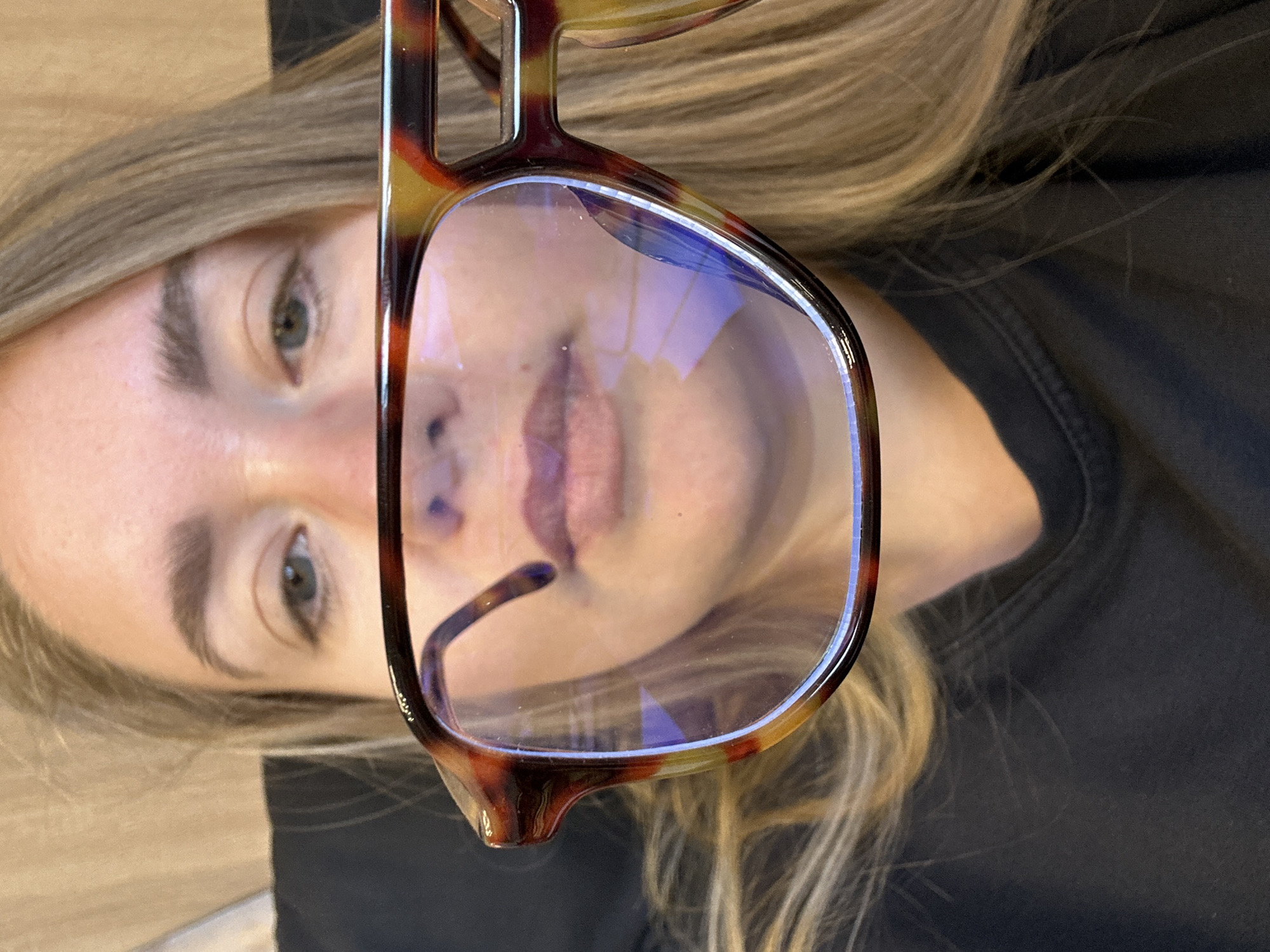 The image shows a person with light blonde hair and fair skin. Their face is partially obscured by a pair of brown and amber patterned eyeglasses. The lenses of the glasses reflect a light source, possibly a window or screen. The person's eyes are visible, appearing to be green or hazel. The background consists of a dark-colored shirt or fabric and a light-colored surface, possibly a wooden floor. The image is rotated counter-clockwise. The location in the city of Ural'skoe, Russia is unknown.