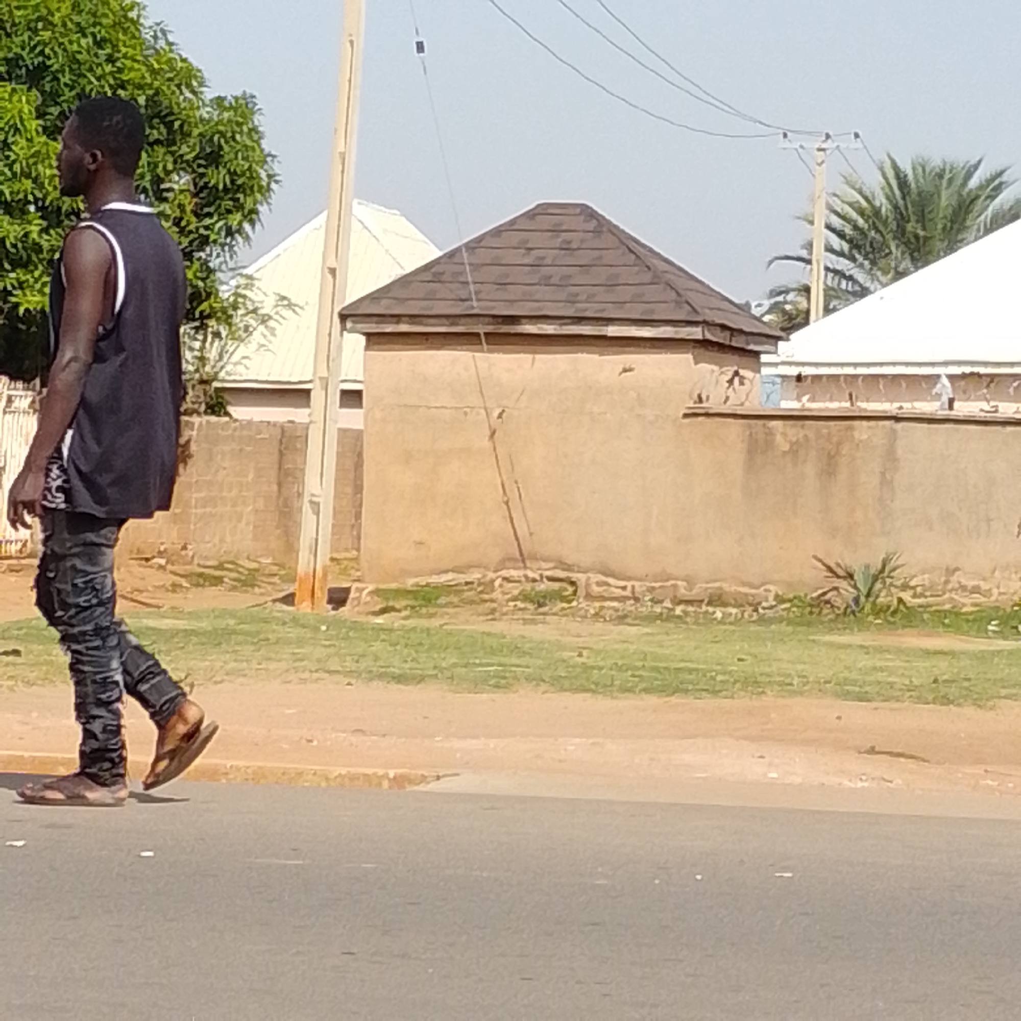 A full-body, side-profile shot shows a young Black man walking on a paved road. He is wearing a black tank top with a white trim, dark grey distressed jeans, and open-toed sandals. He is walking to the left of the frame. In the background, a beige-colored, low-rise building with a dark, pitched roof is visible. A utility pole with wires runs diagonally across the image behind the man and building. Beyond the building, a white-roofed structure and palm trees can be seen. The scene appears to be an outdoor, urban environment, likely in Tempure, Nigeria, with sparse vegetation and a clear sky.
