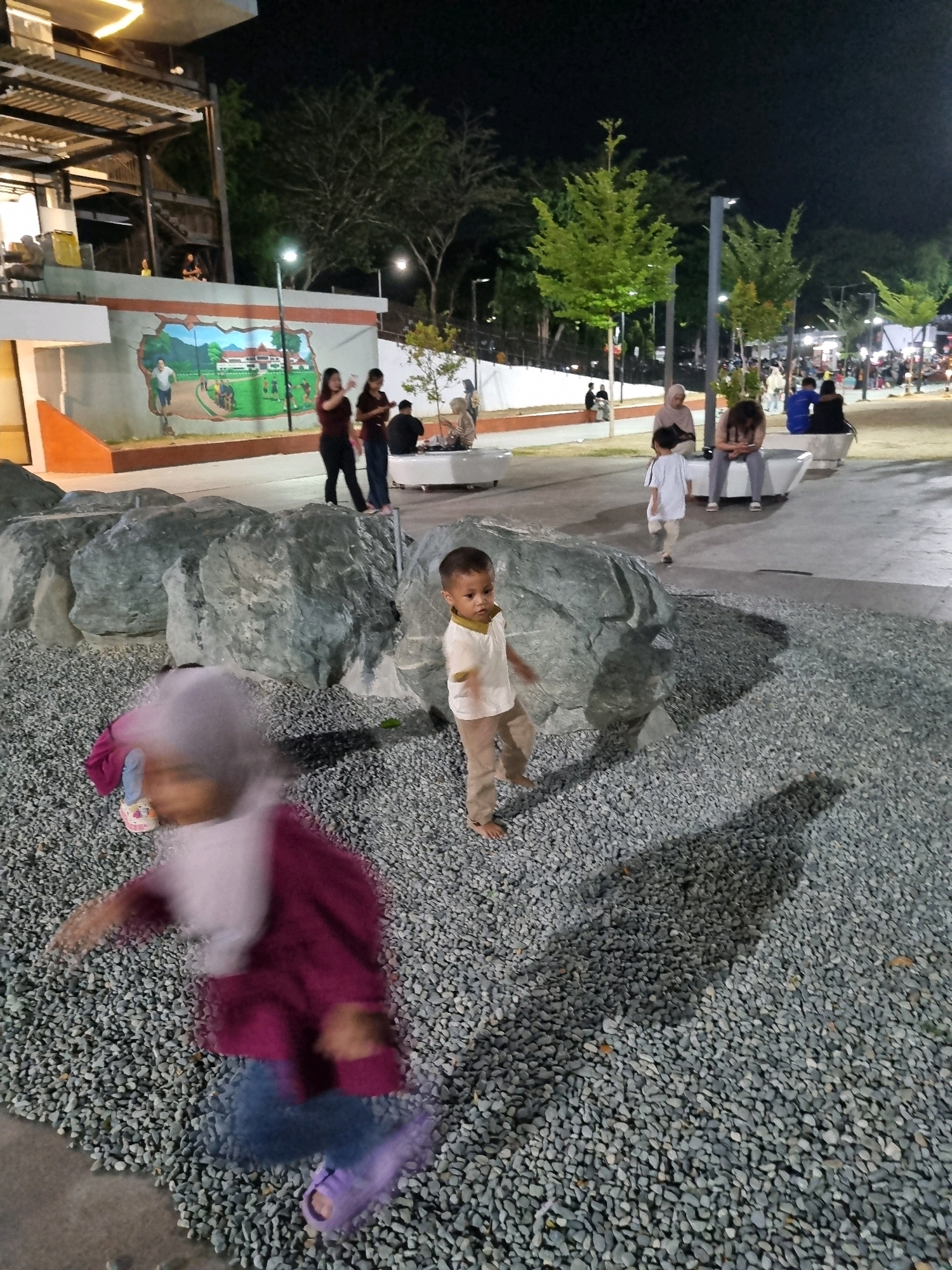 In Tanamodindi, Indonesia, a nighttime outdoor scene unfolds with several people present. In the foreground, a child wearing a purple jacket and blue jeans is captured in motion, their face blurred. To their right, another child in a white shirt and tan pants stands near large rocks. In the background, a group of individuals are seated on white benches, some looking at their phones. Further back, more people are visible, along with trees and illuminated structures, suggesting a public gathering space or park. The ground is covered with small grey stones.