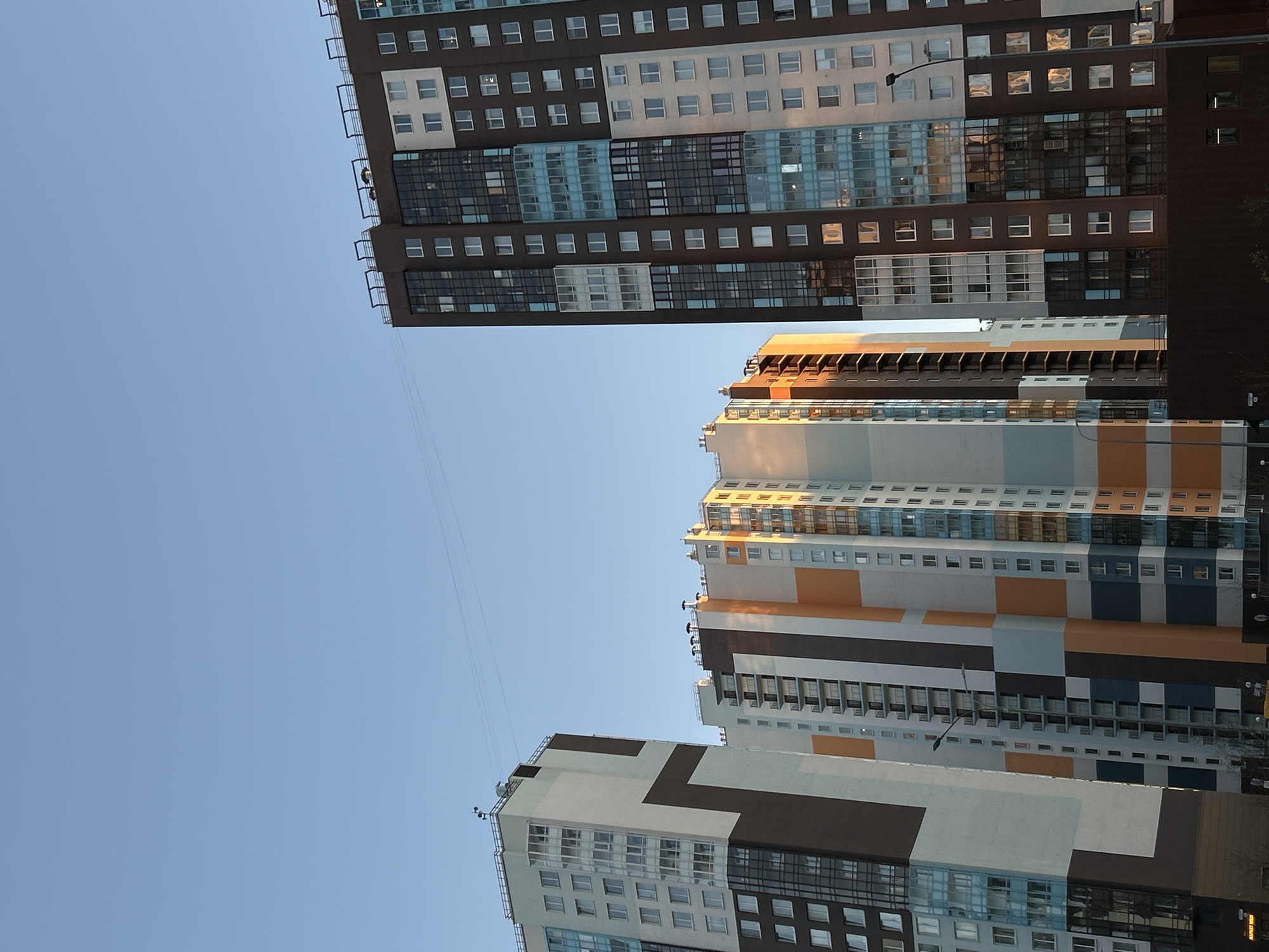 The image depicts a low-angle view of several modern apartment buildings in what appears to be the city of Saint Petersburg, Russia. The buildings feature a mix of architectural styles and color palettes, with combinations of white, gray, brown, and orange hues. The facades are marked by numerous windows. The sky is clear and blue, suggesting a bright, sunny day. A power line runs diagonally across the frame.