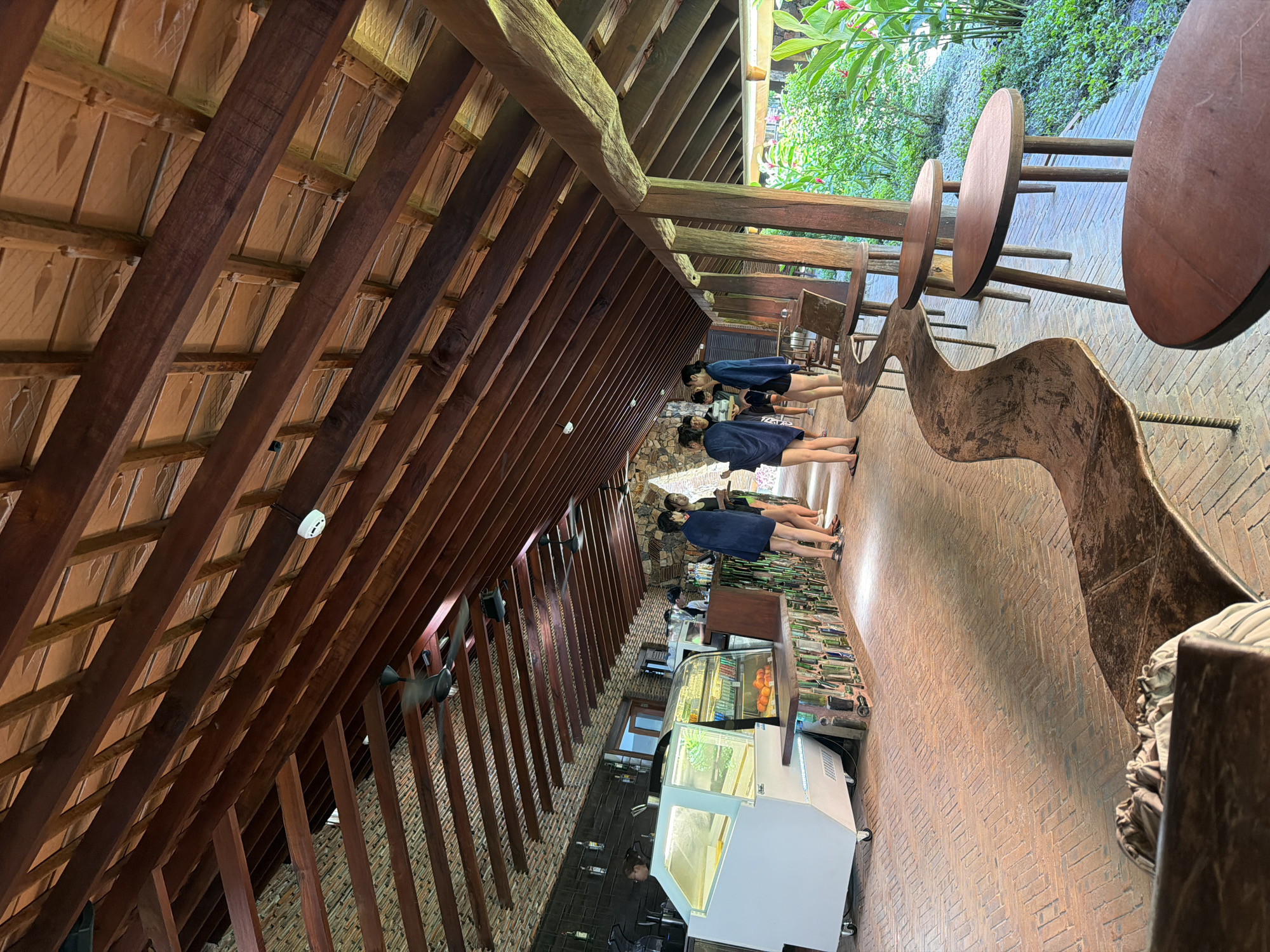 The image depicts a covered outdoor seating area adjacent to a counter service establishment, likely a cafe or bar, located in the city of Tay Nha Trang Ward, Vietnam. The architecture features a prominent wooden beam ceiling with a sloped design, supported by rustic wooden pillars. The floor is paved with reddish-brown bricks laid in a herringbone pattern.
In the foreground, several round wooden tables with three legs are arranged along the right side. A long, winding, dark brown wooden bench with a textured surface is positioned along the left side, running parallel to the counter area.
The counter service area on the left is illuminated by a refrigerated display case stocked with food items, including oranges. Behind the counter, a brick wall serves as the backdrop. A person is visible behind the counter, attending to the service.
Several individuals, appearing to be guests, are standing near the counter, some are holding towels or blankets over their shoulders, suggesting they might be transitioning from or to an outdoor activity like swimming. They are dressed casually, with shorts and some wearing sandals.
The environment to the right of the seating area is lush with green foliage and tropical plants, with hints of red flowers visible. The overall scene conveys a relaxed, tropical atmosphere characteristic of a coastal resort or cafe.