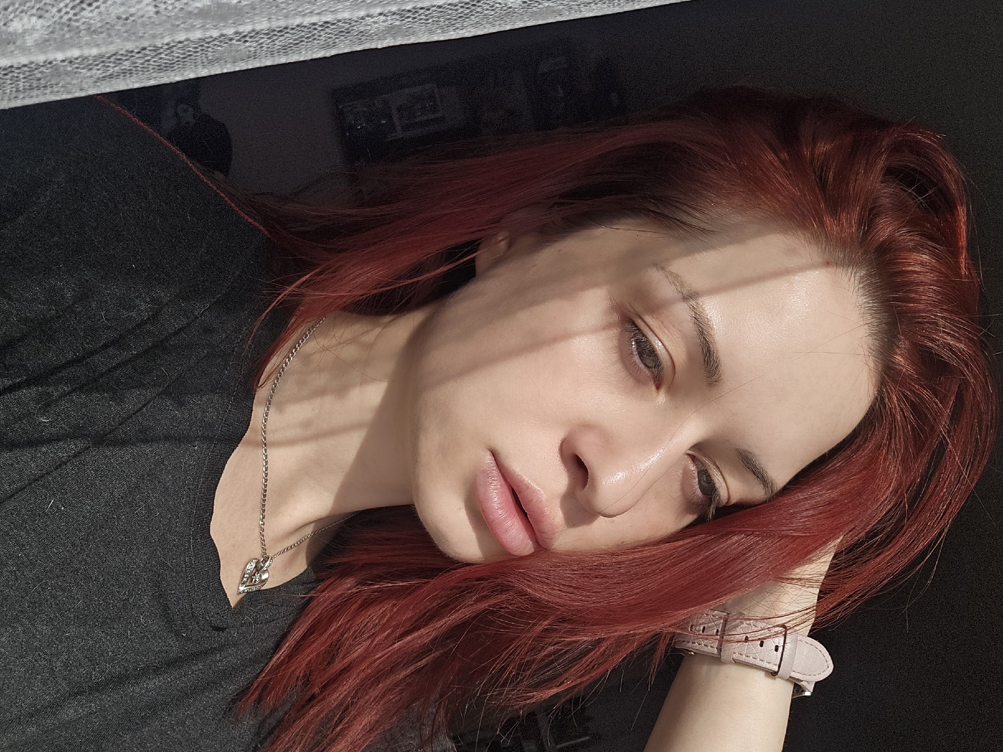 A woman with red hair lies with her head resting on her hand. She is wearing a dark grey shirt and a silver necklace with a heart-shaped pendant. A pink watch is on her wrist. The scene is lit by natural light. The location in the city of Burgas, Bulgaria is not discernible from the image.