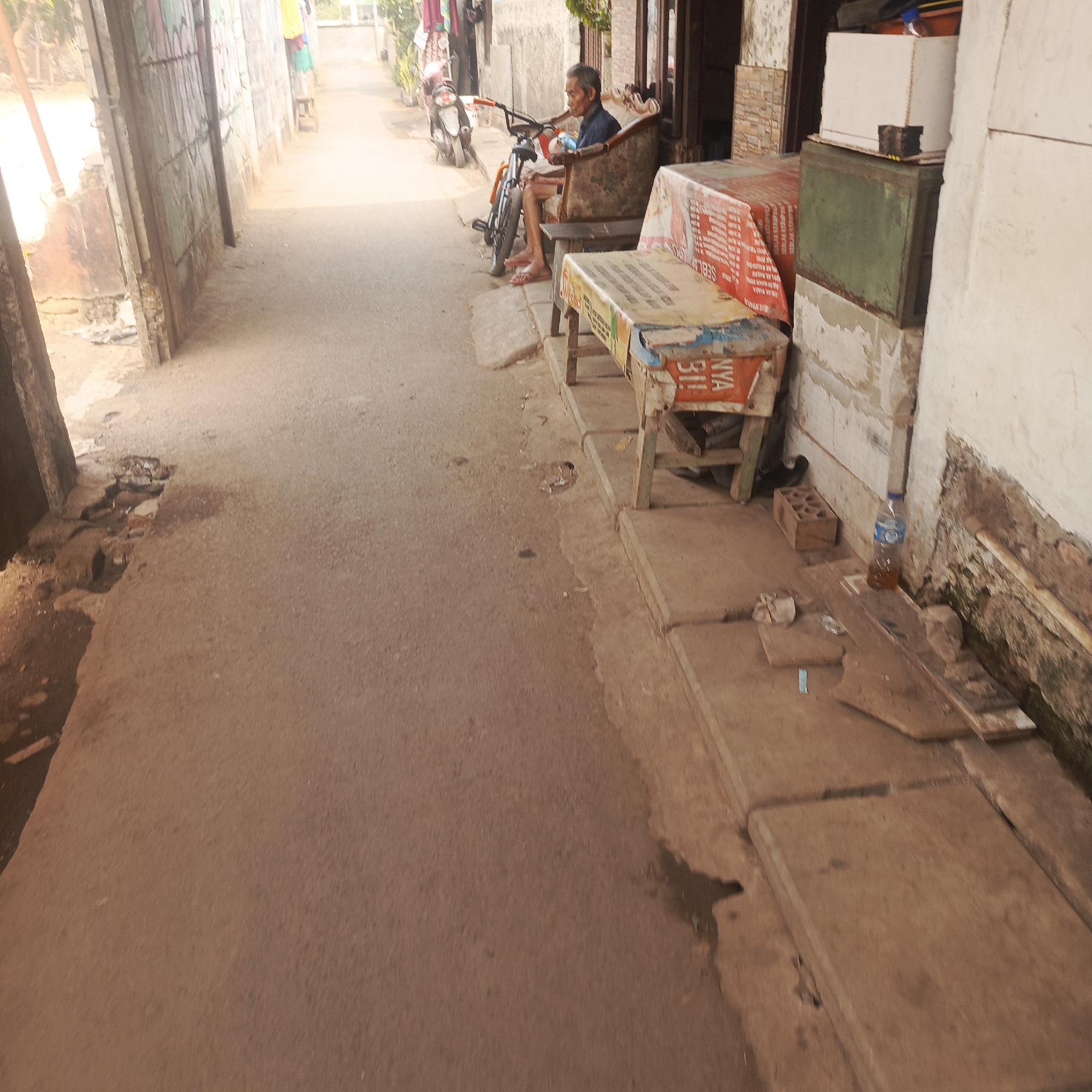 The image captures a street scene in South Jakarta, Indonesia. In the foreground, a narrow, sun-drenched asphalt path leads into the distance, bordered on the left by a weathered, grey stone wall and an open doorway. On the right, a concrete sidewalk runs alongside the path.
Further down the path, a man sits on a patterned armchair, facing a bicycle parked beside him. He appears to be an older gentleman with grey hair, wearing a blue shirt and pink sandals. Beside the man, a wooden table is covered with a red and white patterned cloth. To the right of the table, a green metal chest sits atop a white box. A plastic bottle of water rests on the sidewalk near a brick.
The environment suggests a residential or commercial area with a slightly unkempt appearance, characterized by the cracked pavement and scattered debris. The overall lighting indicates it is daytime, with a strong presence of sunlight casting shadows.