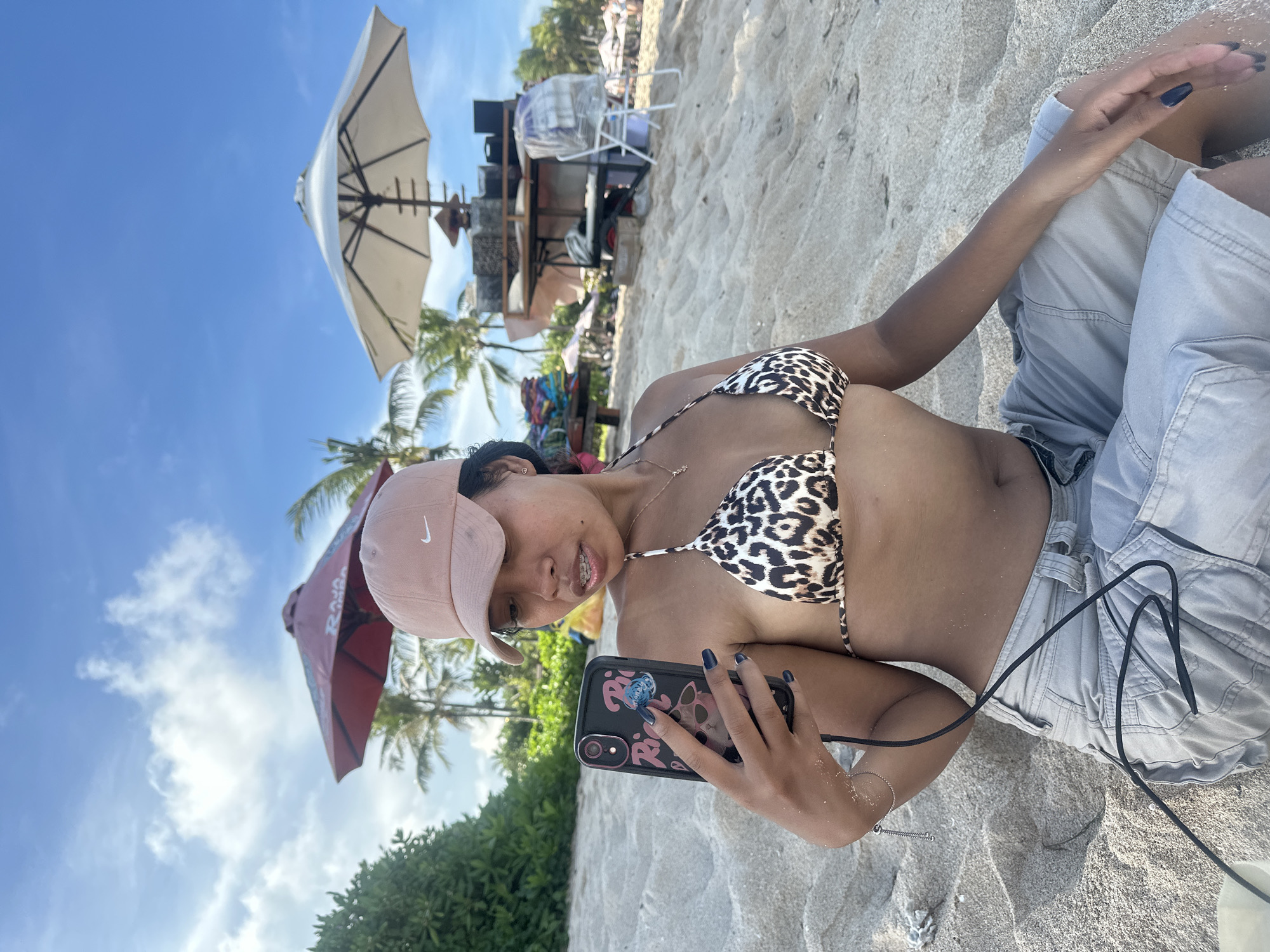 A young woman is shown relaxing on a sandy beach under a bright blue sky with scattered white clouds. She is lying on her side, angled towards the viewer, wearing a leopard print bikini top, light grey shorts, and a light pink baseball cap with a white Nike logo. Her dark hair is mostly covered by the cap, and she has braces on her teeth, accompanied by a slight smile. Her fingernails are painted dark blue.
She is holding a black smartphone with a decorative pop socket or grip that has pink and blue elements. The phone case features a graphic design that includes what appears to be stylized text and a cartoon figure. A black cable is visible, connected to her phone and extending across her body.
The background reveals a typical beach scene with fine sand. Several large beach umbrellas are scattered across the area, including a prominent white one and a darker, possibly red or maroon, one. Palm trees and lush green foliage indicate a tropical setting. Further back, there are glimpses of beach chairs and what appears to be a small vendor stand or cart. The sunny daylight suggests a pleasant day for beach activities. The woman appears to be enjoying her time at the beach, possibly taking a selfie or recording content with her phone. The setting aligns with a tropical beach environment like Kuta Selatan, Indonesia.