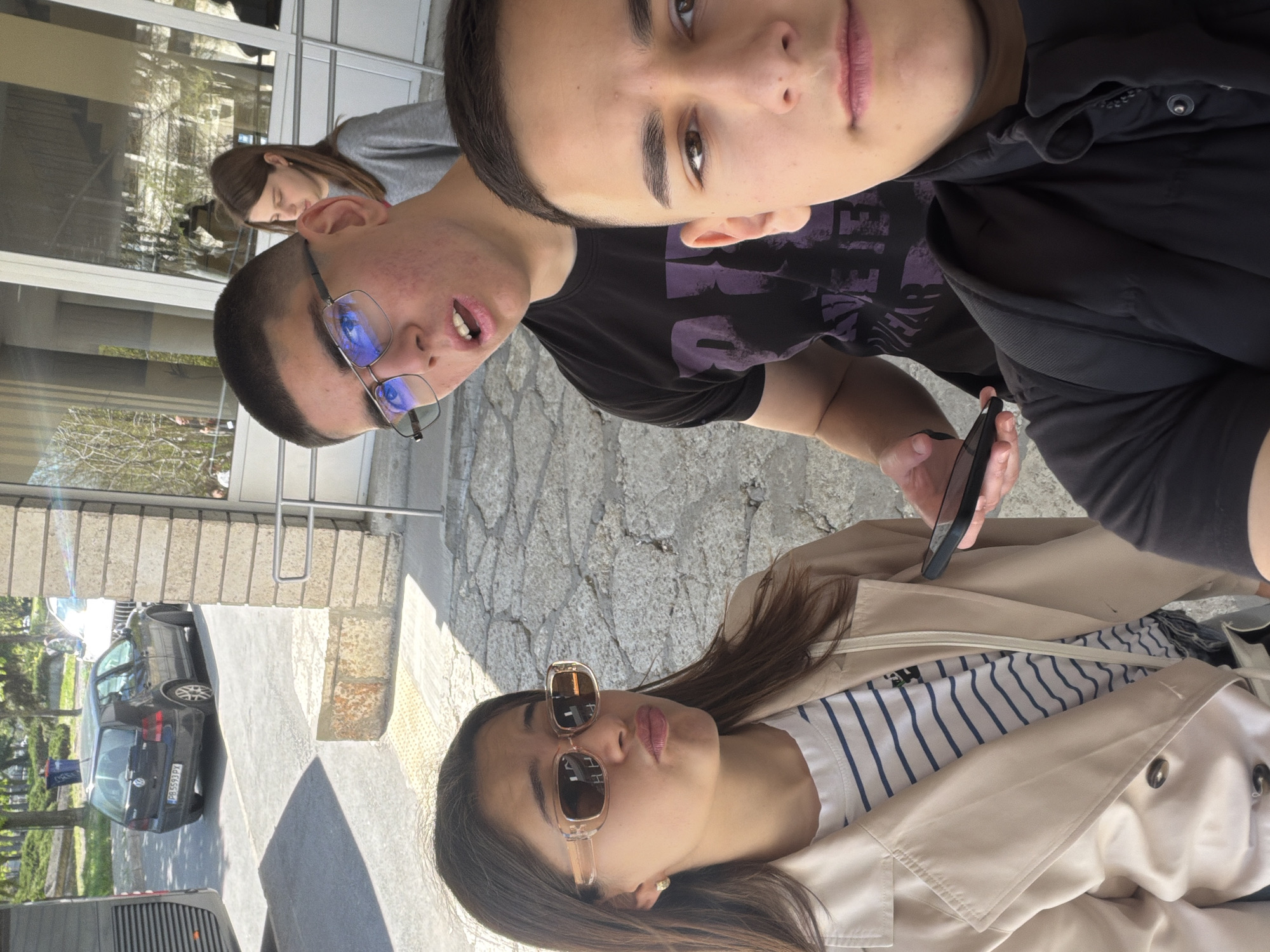 The image depicts three young people in an outdoor urban setting in Shumen, Bulgaria, likely posing for a casual group photo or selfie during the daytime. The lighting suggests a bright, sunny day, casting discernible shadows.
In the foreground, a young woman with long dark hair is prominent, wearing sunglasses, a white t-shirt with thin blue horizontal stripes, and a light beige trench coat. She looks towards the camera with a slight pout. Behind her, and slightly to the right, a young man with short dark hair gazes directly at the camera with a neutral expression. He wears a black t-shirt featuring a faded purple graphic, where partial text such as "EHE" is visible.
To his left and slightly in the background, another young man is seen. He wears eyeglasses that reflect blue light, has short dark hair, and visible facial blemishes. His mouth is slightly open, and he is holding a black smartphone in his right hand, potentially using it to capture the image. Reflected in a large glass window behind them, a fourth person, appearing to be a young woman with long brown hair and a gray top, is partially visible.
The scene includes stone-paved ground, concrete structures, and metal railings. A dark-colored Volkswagen Golf station wagon is parked in the background, displaying the license plate "PB 5593 PX". Part of another vehicle is also visible in the lower left corner. The overall atmosphere is relaxed, with the subjects appearing comfortable in their outdoor environment.