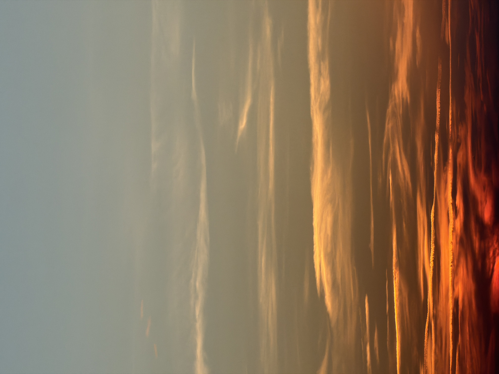 The image shows a portion of the sky with a gradient transitioning from a light, muted gray-blue on the left to a deep, fiery orange-red on the right. The transition occurs across elongated, vertical cloud formations. The leftmost area is mostly clear, while clouds become increasingly prominent toward the right, displaying vertical striations that are highlighted by the sun's glow. The clouds closer to the right are densely packed and have a more pronounced, turbulent texture. The location details are not provided.