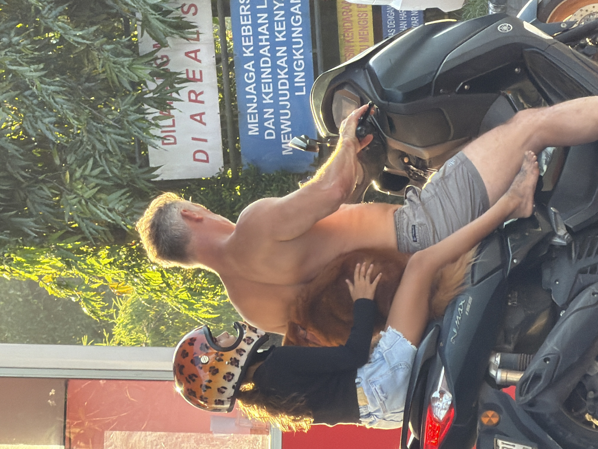 The image shows a shirtless man on a Yamaha NMAX scooter in Mengwi, Indonesia, with a young girl wearing a leopard print helmet sitting in front of him. A sign partially visible in the background on the left reads "DILKA DIAREALIS". A sign in Bahasa Indonesian is partially visible behind the man, translating roughly to "Maintain cleanliness and beauty of the environment to realize comfort in the environment".