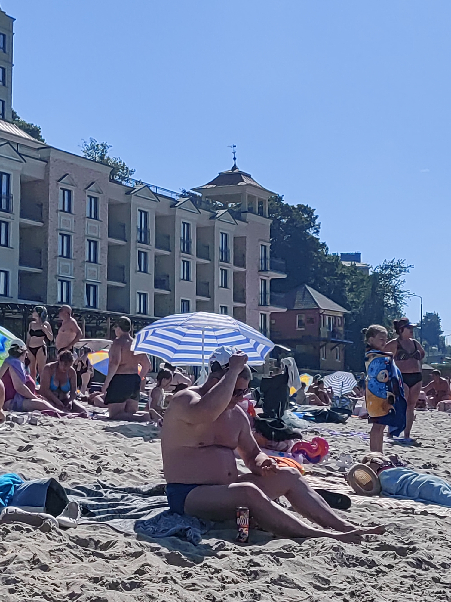 In Pionersky, Russia, on a sunny day, a crowded beach is depicted. Several people are seen sunbathing and relaxing on the sand. In the foreground, a man is seated on the beach, holding a can of "OXO" drink. Further back, a blue and white striped umbrella provides shade for beachgoers. Several buildings, likely hotels or apartments, are visible in the background, with a clear blue sky overhead. Some individuals are wearing swimwear, while others are covered with towels. The overall scene is one of leisure and activity on a summer day at the beach.