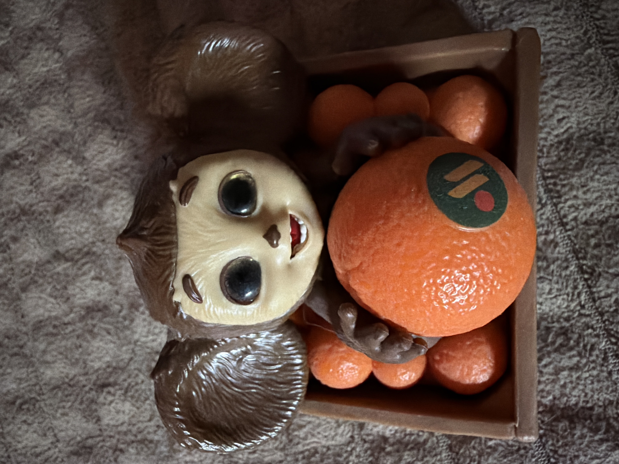 A close-up, top-down shot shows a Cheburashka toy, a popular Russian cartoon character, sitting in a brown plastic crate filled with small, bright orange objects that resemble tangerines. The toy is light brown with large, dark eyes, a wide smile, and big, furry ears. Its paws are positioned around a large, central orange object that also has a sticker with green, orange, and red elements. The crate and the toy are resting on a textured, greyish-brown surface, likely a blanket or carpet. The lighting appears to be overhead, casting subtle shadows and highlighting the textures of the toy and the surrounding surface. The image provides no discernible context or clues to its specific location within the city of Naberezhnye Chelny, Russia.