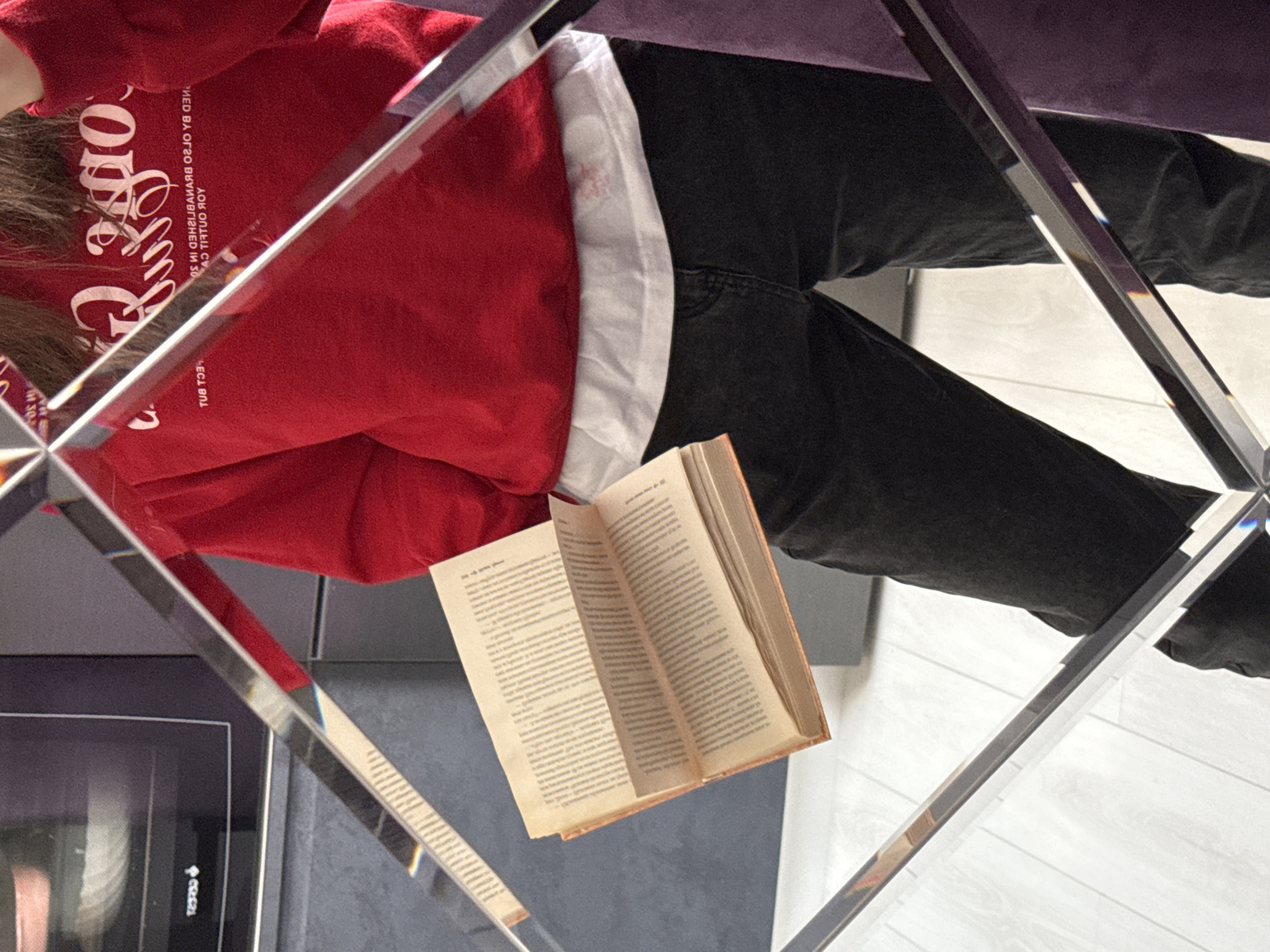 The image shows a person seated, seen through a mirrored surface. The person wears a red sweatshirt with white writing, a white undershirt, and black pants. They are reading an open book. The book is thick and has pages filled with text. The mirrored surface is fragmented, creating multiple reflections of the person and the surrounding environment. The background is indistinct, but appears to be a room with a flat surface, potentially a floor or tabletop. A portion of an electronic device is visible in the lower left corner. The location of the scene is unknown.