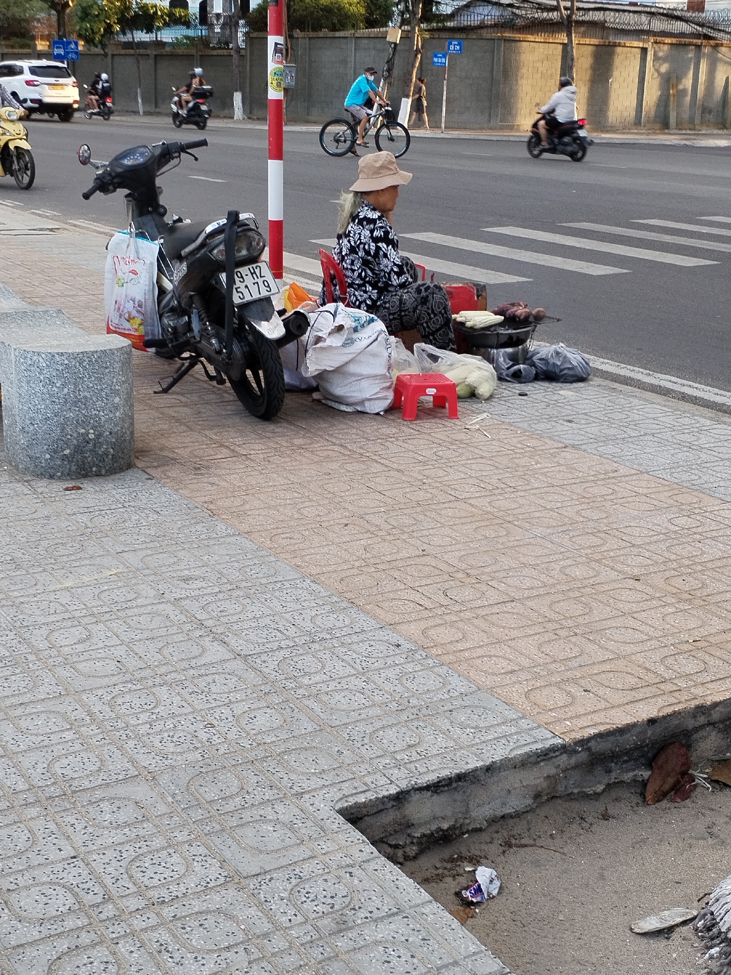 The image depicts a street scene in Bac Nha Trang Ward, Vietnam. A woman wearing a hat and floral print clothing is sitting on a red plastic stool on the sidewalk next to the road. She appears to be selling grilled corn and other items from a small grill, with bags of goods and food around her. A black motorcycle is parked nearby, its license plate number is visible: 79-H2 5179.
The street is visible, with a crosswalk and various forms of transportation including bicycles and motorcycles. Other people are present in the background riding their motorbikes on the road. The sidewalk is paved with two different types of concrete tiles, and there is some debris in the gutter between the sidewalk and the road. A red and white striped pole stands next to the sidewalk close to the woman.