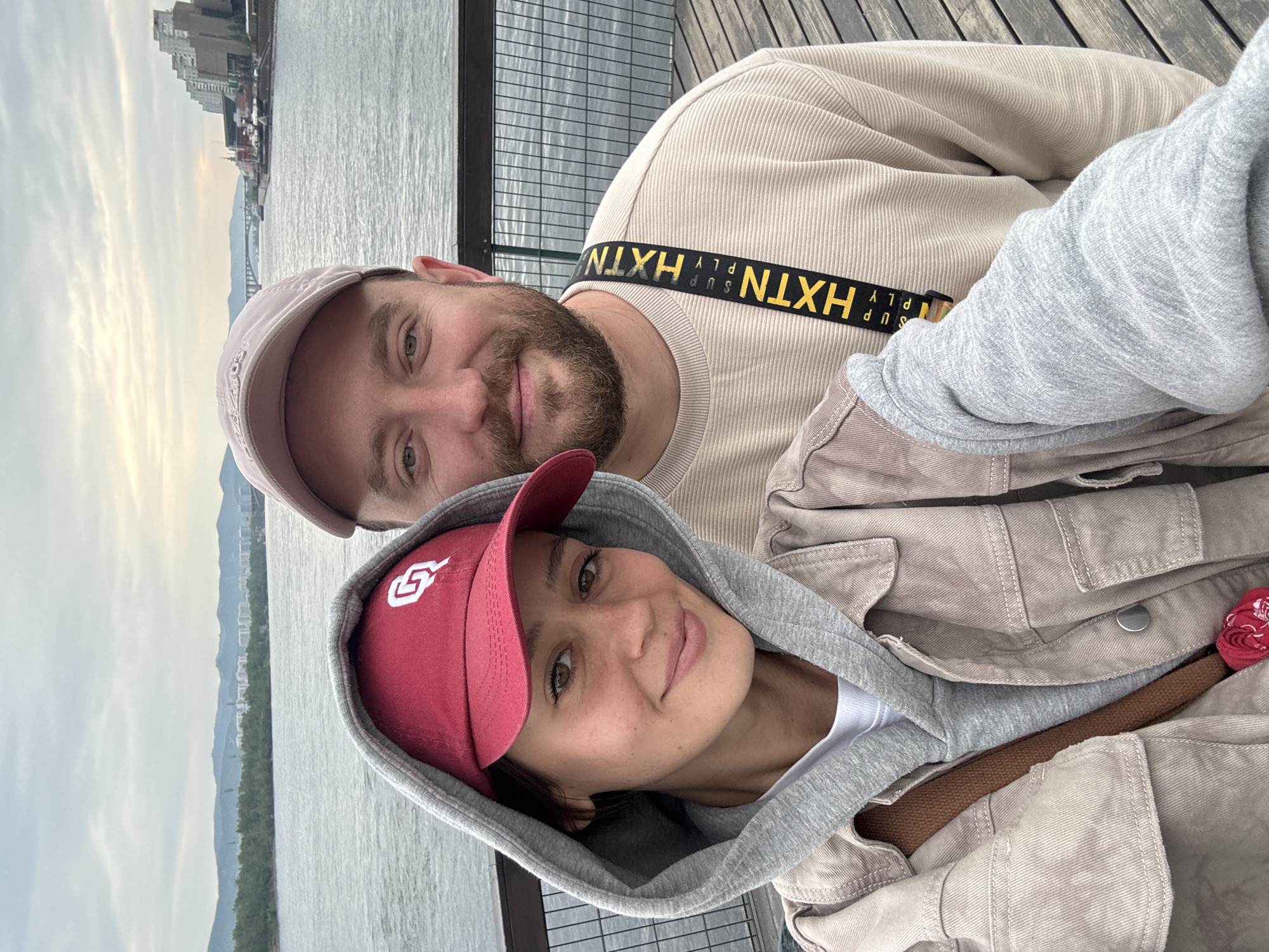 Two individuals, a man and a woman, are taking a selfie on a wooden boardwalk next to a body of water. The woman, on the left, is wearing a grey hooded sweatshirt with the hood up, a tan utility jacket, and a red baseball cap with a white logo. The man, on the right, is wearing a light beige crewneck sweater and a light pink baseball cap. He has a beard and is wearing a yellow strap across his chest with black lettering that reads "HXTN SUPPLY". The background shows a wide river, trees, distant hills, and buildings along the far bank, characteristic of the city of Krasnoyarsk, Russia, specifically near the Yenisei River embankment. The sky is overcast.