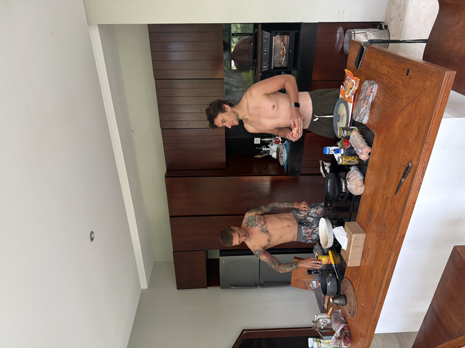 The image depicts an indoor kitchen setting located in Ungasan, Indonesia. Two adult males, both shirtless, are present around a large wooden island counter.
The male on the left, with extensive tattoos covering both arms, shoulders, and chest, wears patterned shorts. He stands over a stovetop integrated into the island, with his left hand positioned above a black pot, next to a white bowl, a small pan, and various spice grinders. A knife rests on the counter. The male on the right, with short dark hair and wearing dark green shorts with a smartwatch on his left wrist, holds a small, reddish object in both hands and faces towards the male on the left.
On the wooden island counter, a gray frying pan, a clear plastic carton of eggs, a loaf of bread in a plastic bag, several canned goods, a blue and white container, and a dark-colored smartphone are visible, along with a tissue box.
The kitchen features dark wood floor-to-ceiling cabinetry along the back wall. To the left of the cabinetry, a silver refrigerator is present. To the right, above a black countertop, a toaster oven contains food, and a reflection of outdoor foliage is visible in the surface behind it. The ceiling is white with a single recessed light fixture. The walls are light-colored, and a wooden bar stool is partially visible in the lower-left foreground.