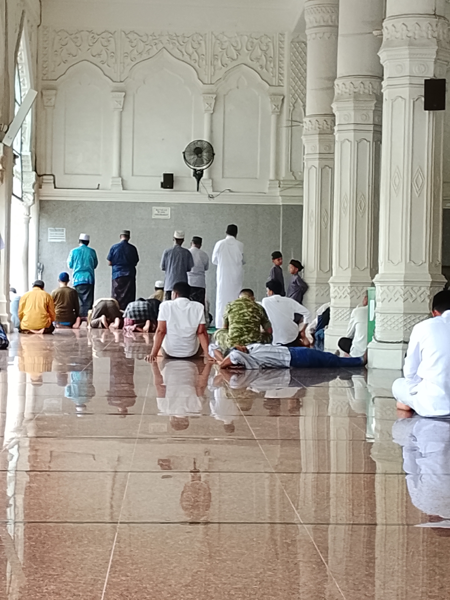 The image depicts a group of men engaged in prayer within a mosque. The scene is set in an indoor space characterized by polished, reflective flooring, tall, decorative pillars, and white, ornate wall carvings. Several men are visible in various stages of prayer, some standing, some kneeling, and others prostrate on the floor. The men are dressed in traditional Islamic attire, including caps and long robes. The lighting suggests it is daytime, possibly with natural light filtering in from unseen openings. The architectural style points towards a Southeast Asian Islamic setting, and the presence of a ceiling fan suggests a warm climate. The specific location is the Baiturrahman Grand Mosque in Lhokseumawe, Indonesia, as indicated by the architectural style consistent with prominent mosques in the region and the typical cultural practices observed.