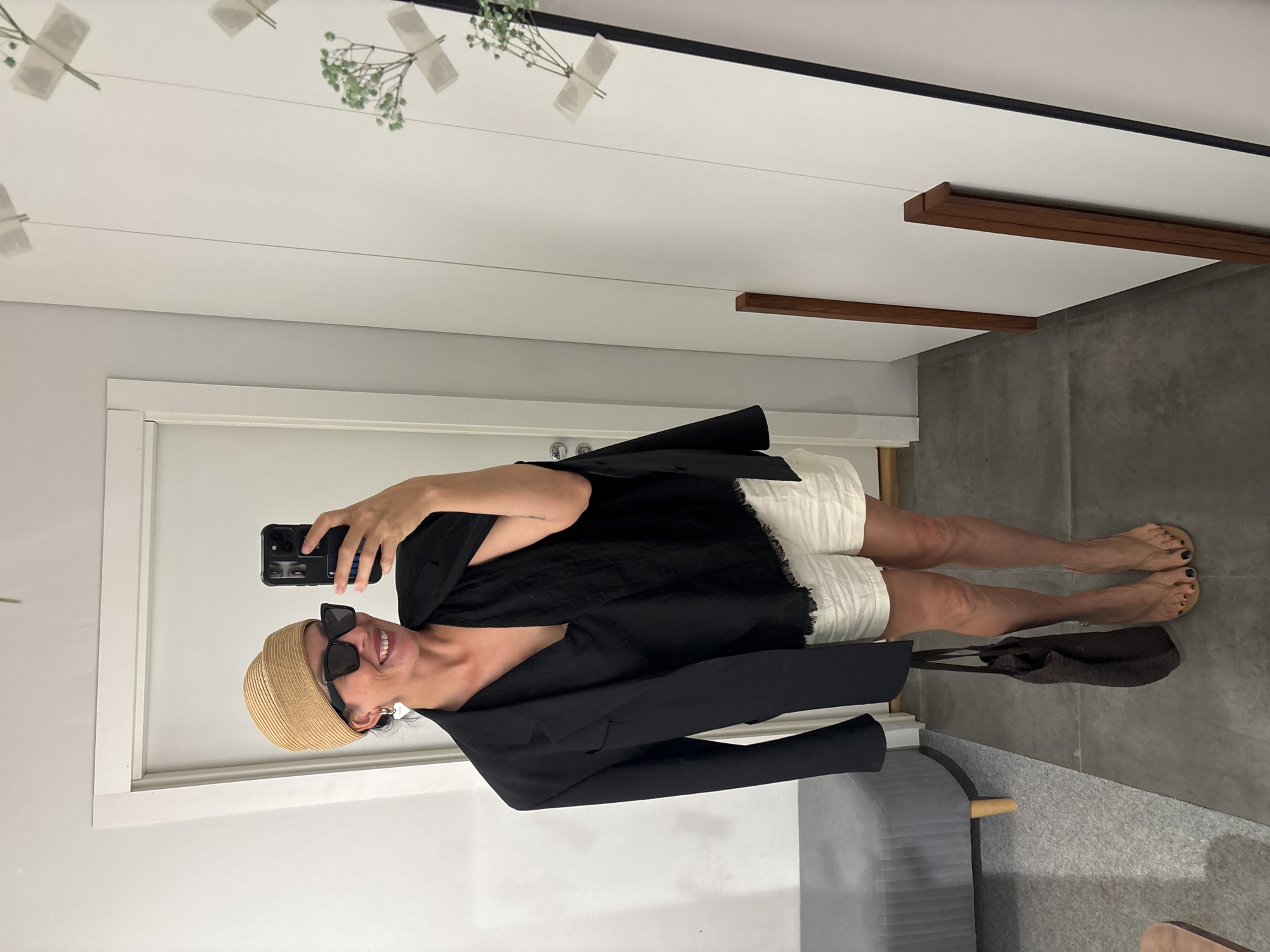 A woman stands indoors, posing for a mirror selfie. She is wearing a beige woven hat, black sunglasses, a black blazer draped over her shoulders, a black V-neck top with frayed edges, and white linen shorts. She is also holding a dark brown tote bag and wearing tan flip-flops. The environment appears to be a dressing room or a room with a mirror. There is a grey upholstered bench to the left of the frame and a white closet or wall with wooden trim and decorative dried flowers on the right. The floor is a smooth, grey concrete. The image does not provide any information about the location in the city of Chaikovsky, Russia.