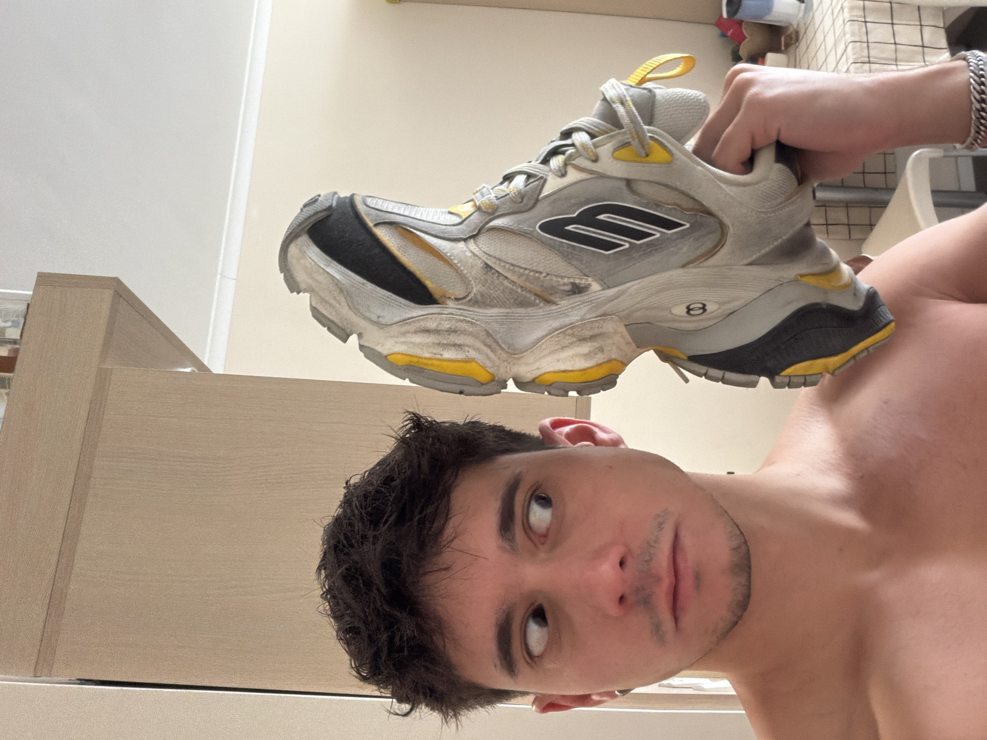 A medium shot presents a shirtless man holding a Balenciaga Runner sneaker overhead. The man is Caucasian, with dark hair, light eyes, and sparse facial hair. He gazes upward toward the shoe. The shoe itself is a gray, yellow, and black sneaker held in the right hand of another individual, visible by the wrist and a silver chain bracelet. The background shows a cream-colored wall. The location is not identifiable as Sofia, Bulgaria, based solely on this image.
