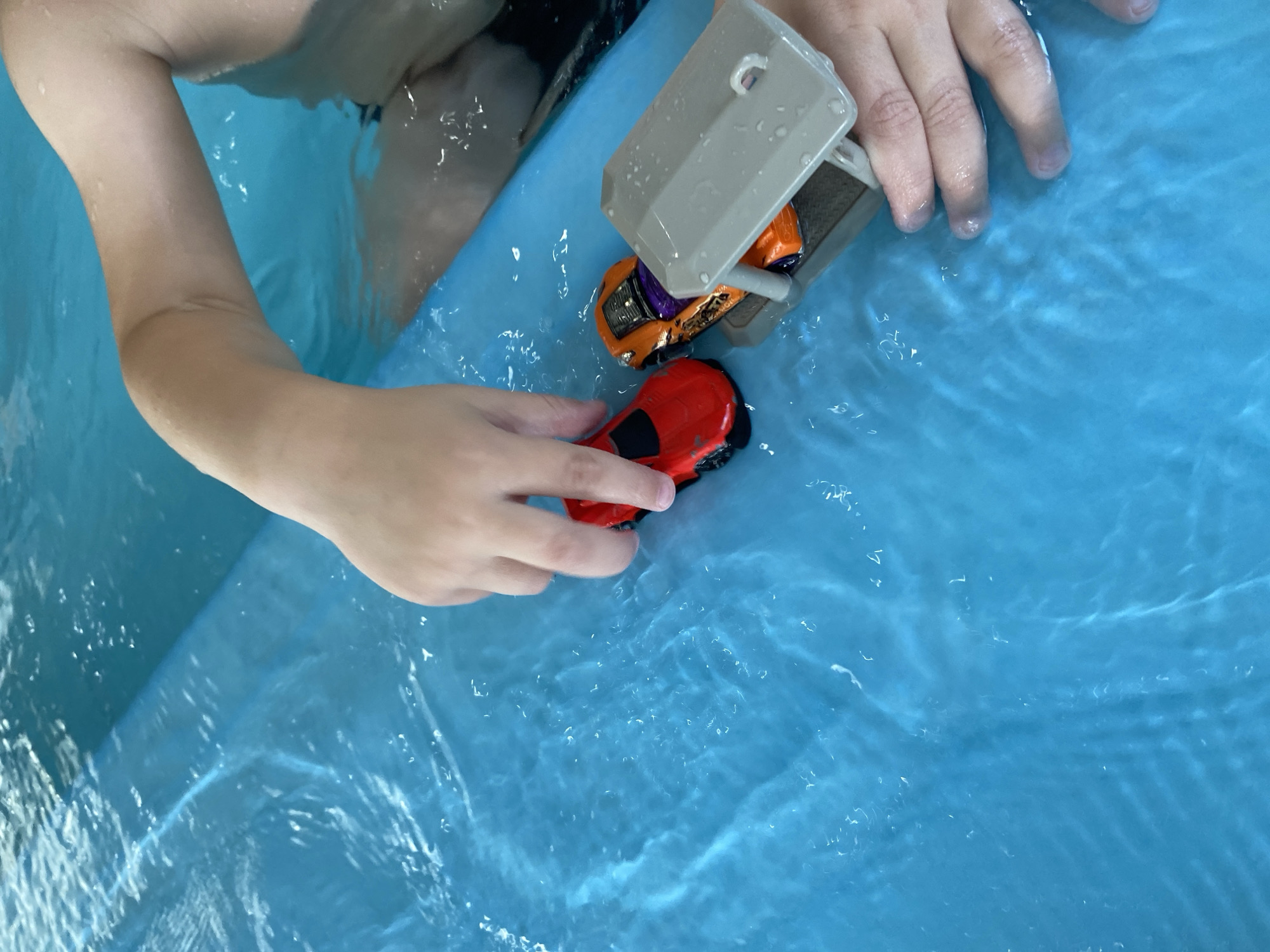 The image shows a child's hands playing with toy cars in a bright blue pool. A red toy car is to the left, and an orange and purple toy car is positioned under a gray plastic structure to the right. The water is clear and rippled, indicating movement. The child's arms are visible, submerged in the water. The background shows the edge of the pool and more water. There is no indication of the location being in Chulis, Russia.